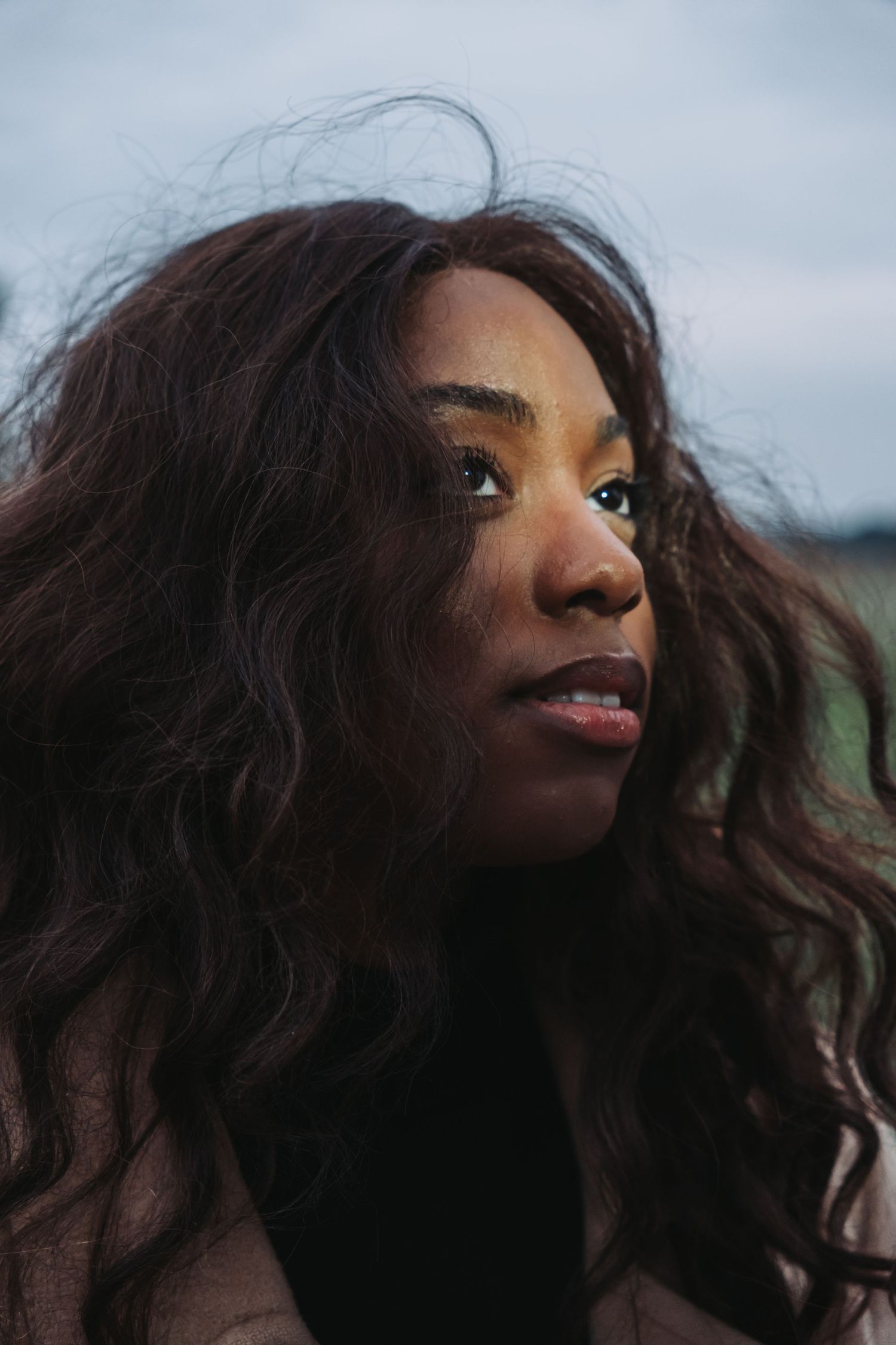A young Black woman with long wavy dark hair gazes thoughtfully into the distance outdoors on a cloudy day.