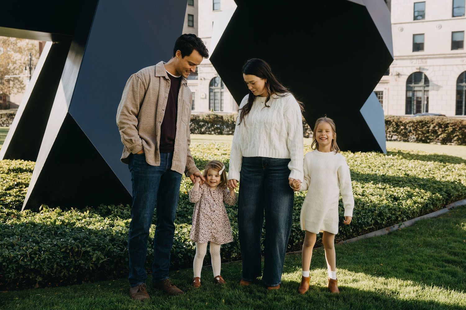 Family of four holding hands outdoors near modern black sculpture and green hedges on a sunny day.