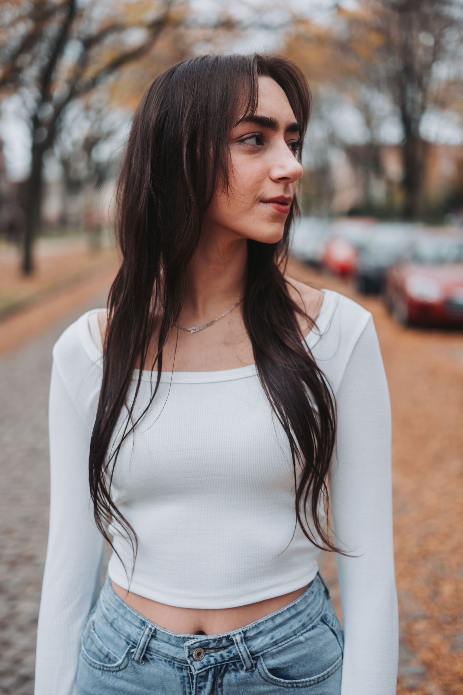 Young woman with long dark hair wearing a white crop top and jeans standing on a tree-lined street.