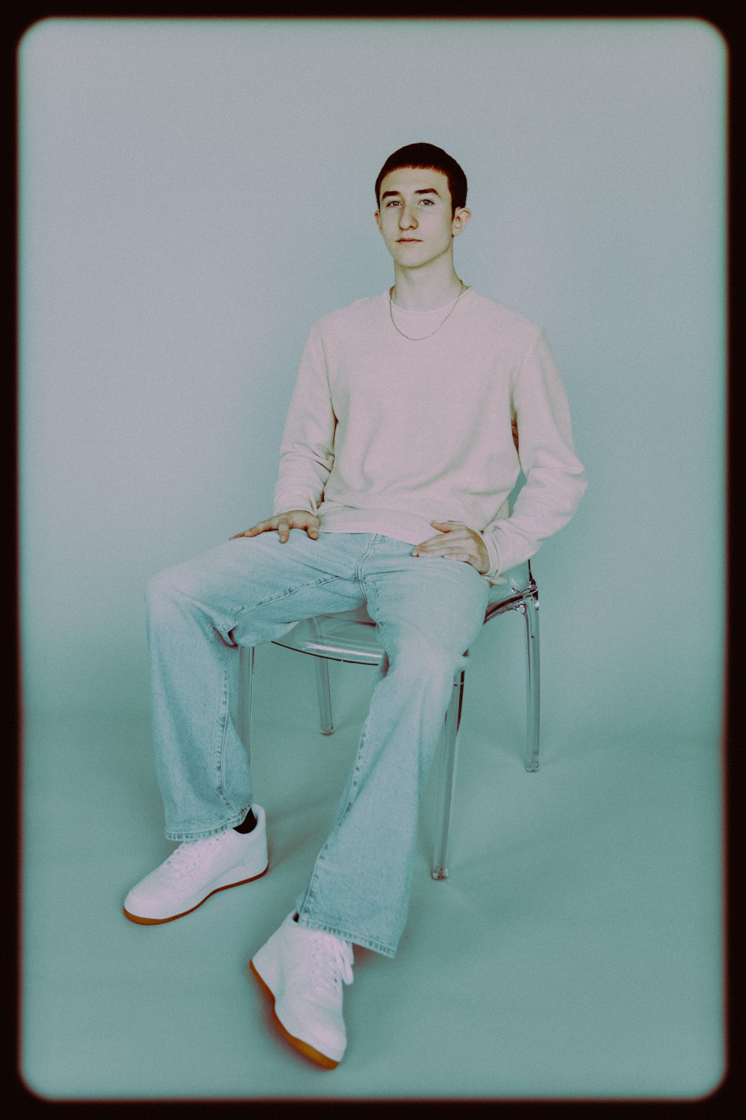 Young man in pink sweater and wide-leg jeans sits on metal chair against mint background wearing white sneakers.
