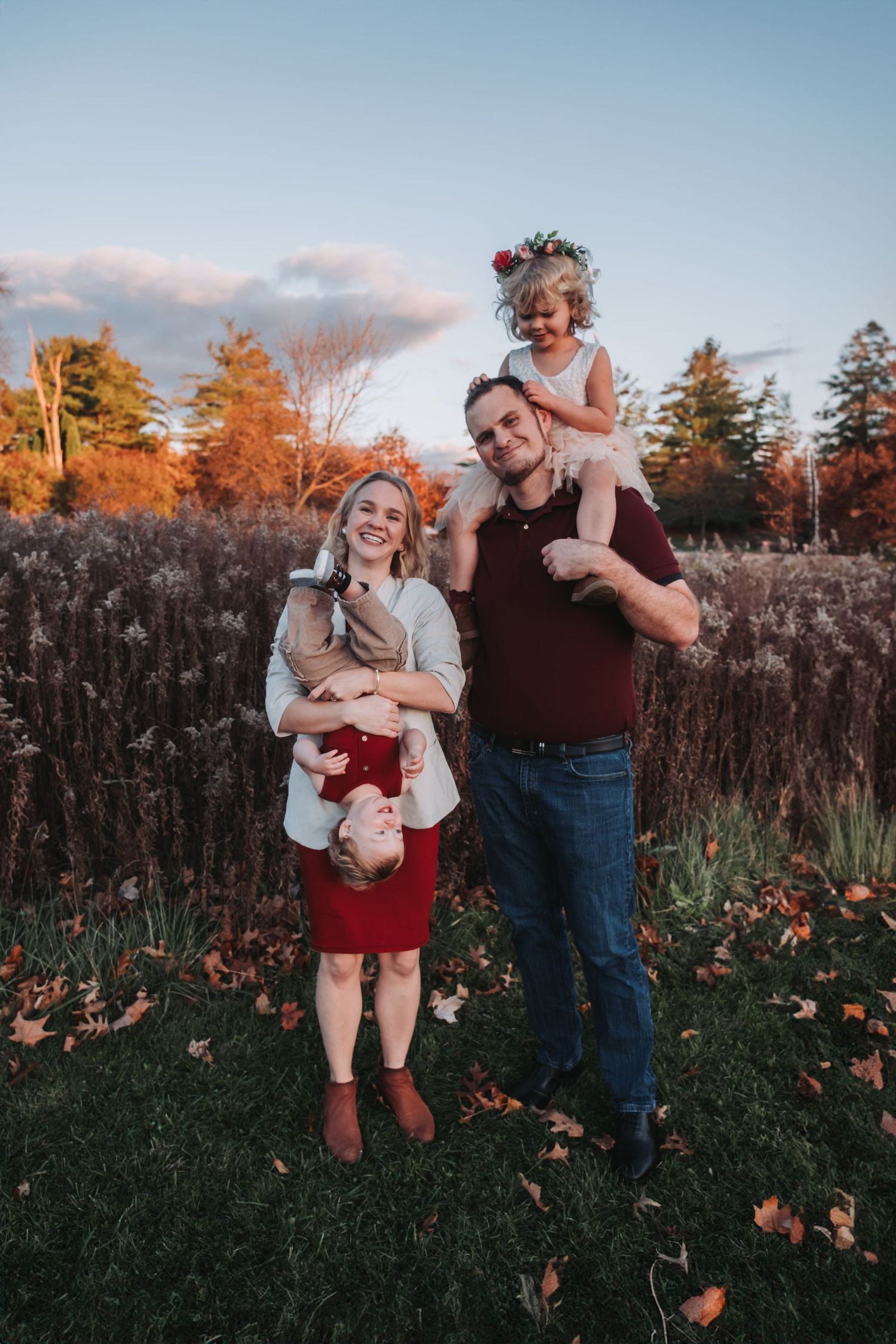 Family of four poses outdoors in fall, mom holding baby upside down, dad with toddler on shoulders, autumn foliage background.