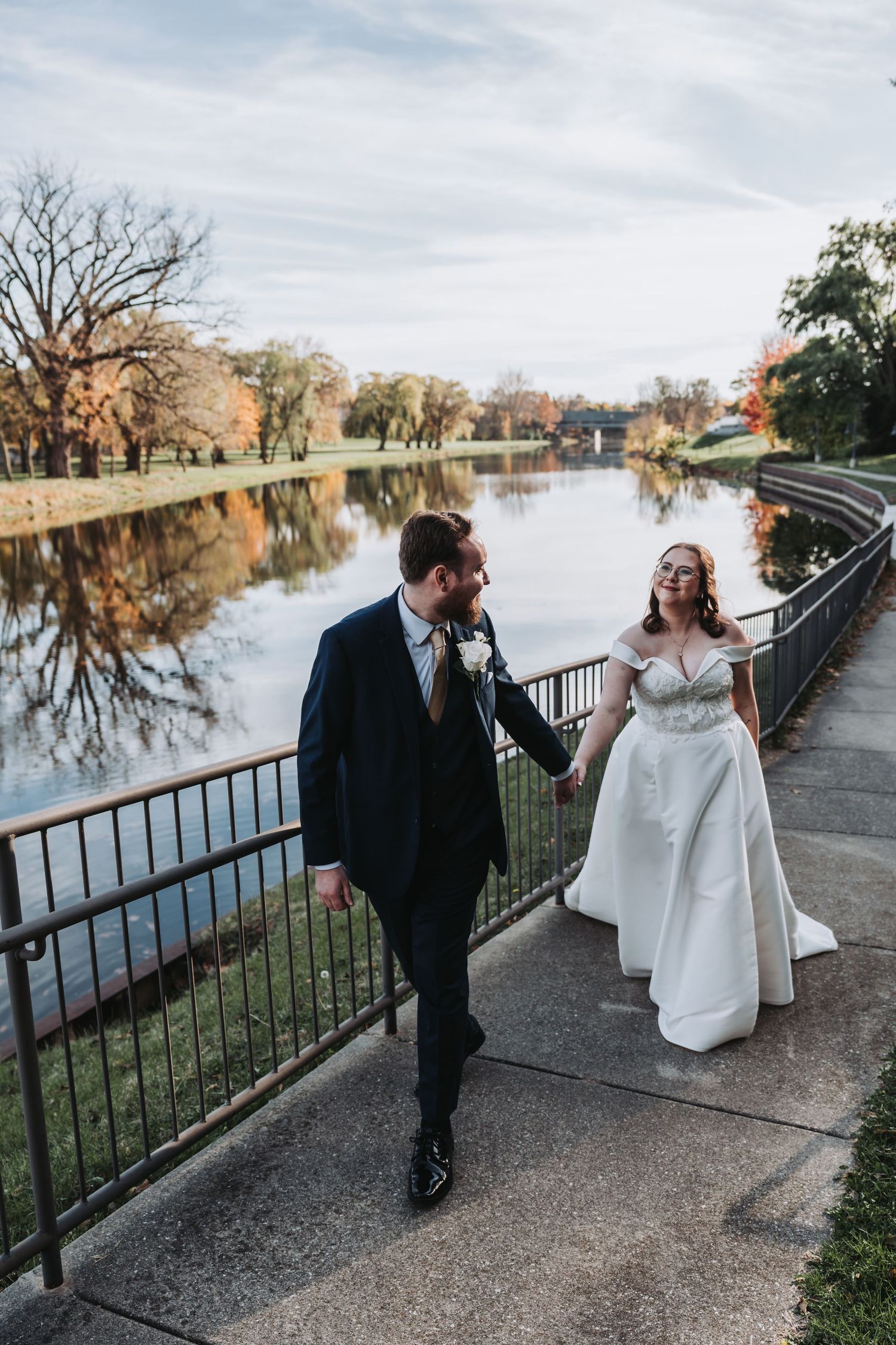 Bride and groom walk hand in hand along scenic riverside path with fall foliage reflections.