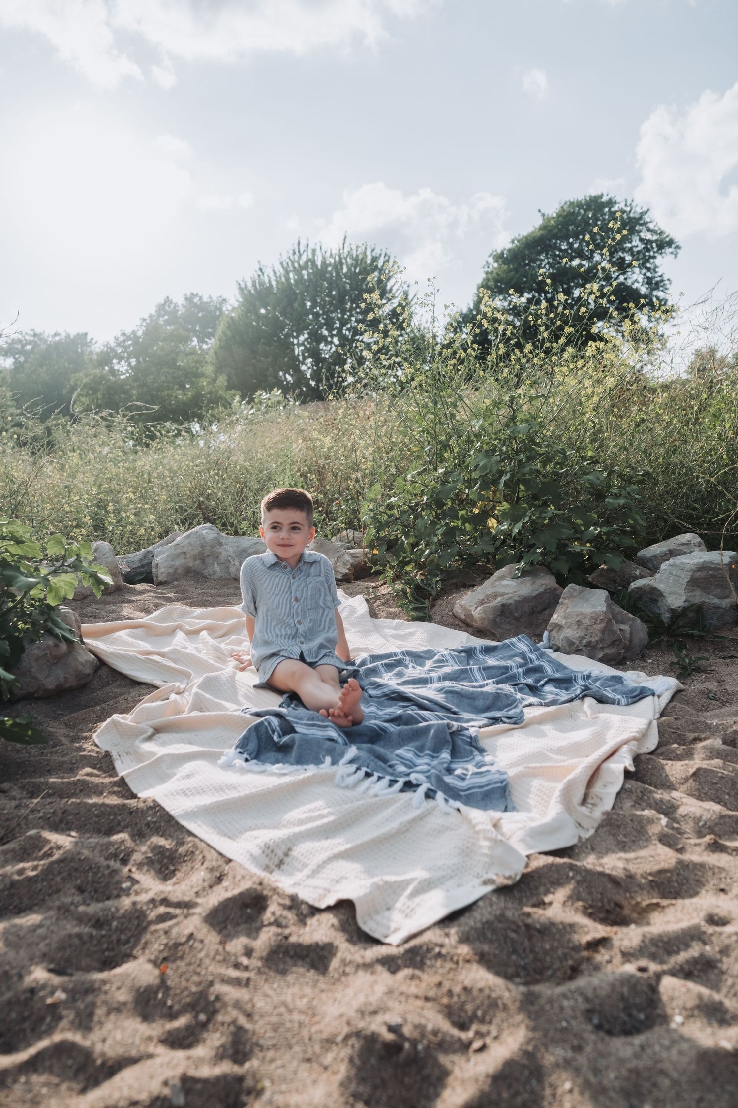 Young boy sitting on a blanket on sandy beach surrounded by rocks and green trees on a sunny day.