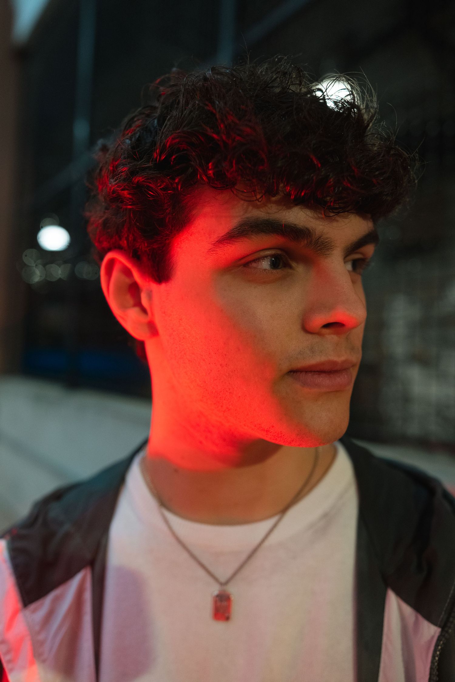 Young man with curly hair lit by red neon light at night, wearing a white tee and jacket with a pendant necklace.