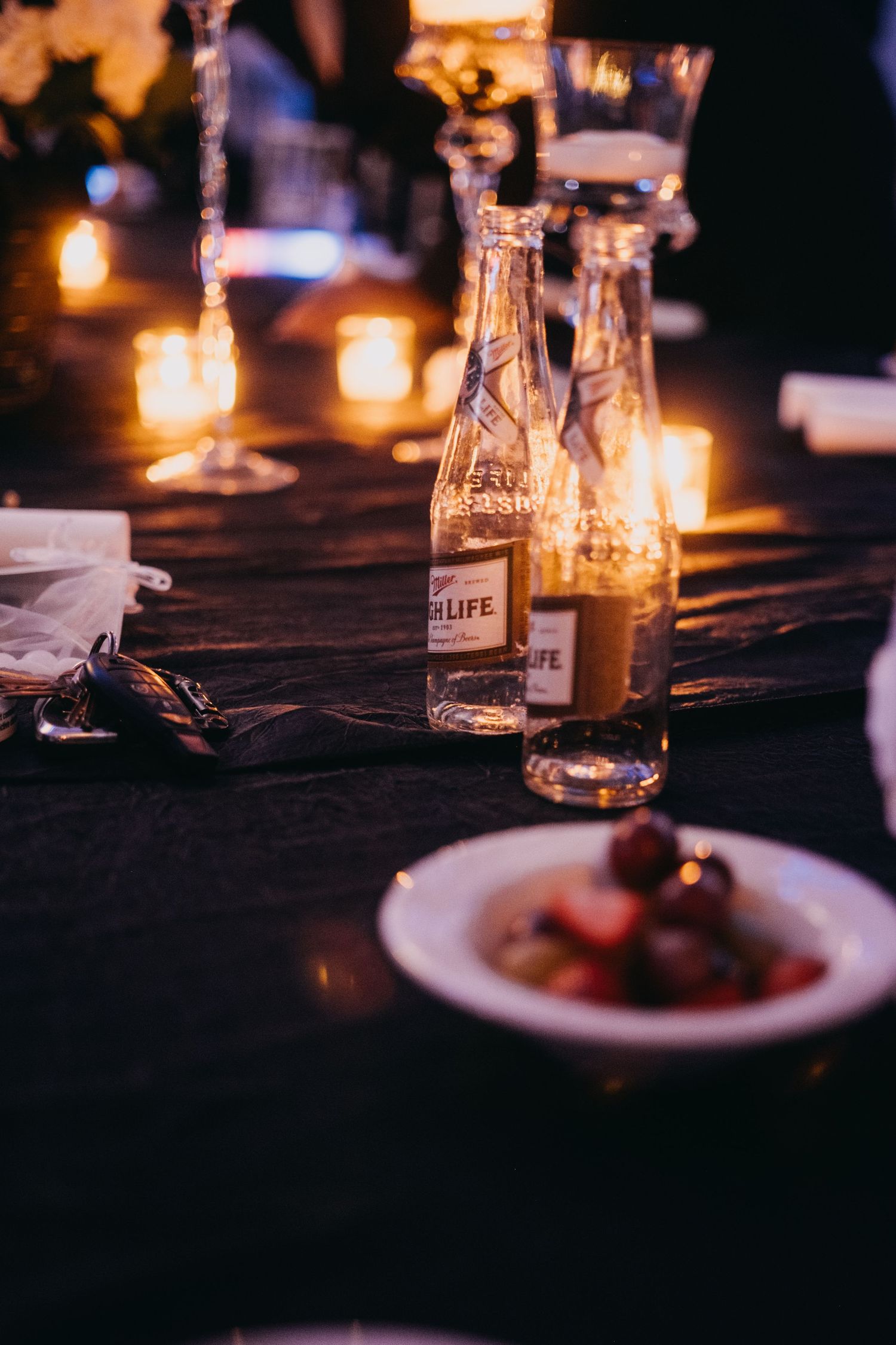 Candlelit dinner table with Miller High Life bottles, glowing candles, and a plate of appetizers on dark linen.