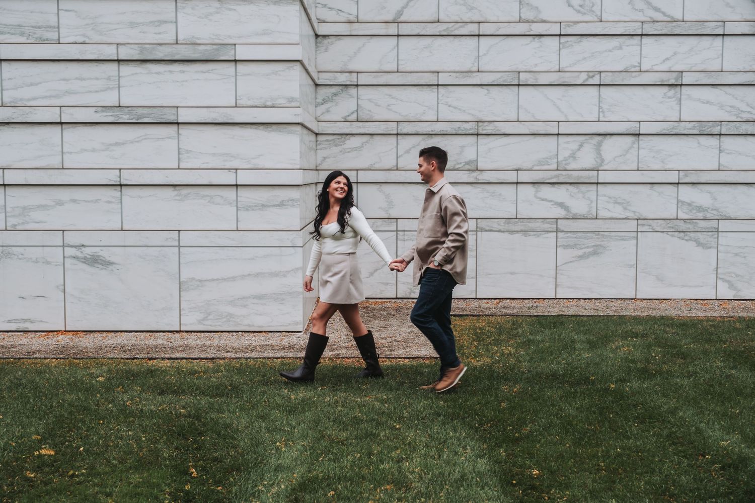 Couple holding hands and walking together laughing in front of a marble wall on green grass.