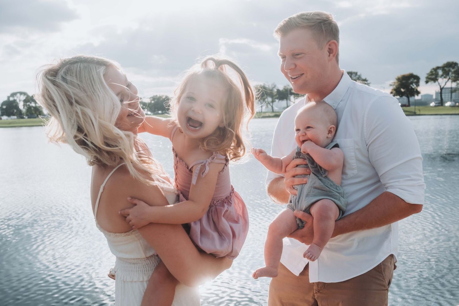 Happy family of four laughing outdoors near a lake, parents holding two young children on a cloudy day.