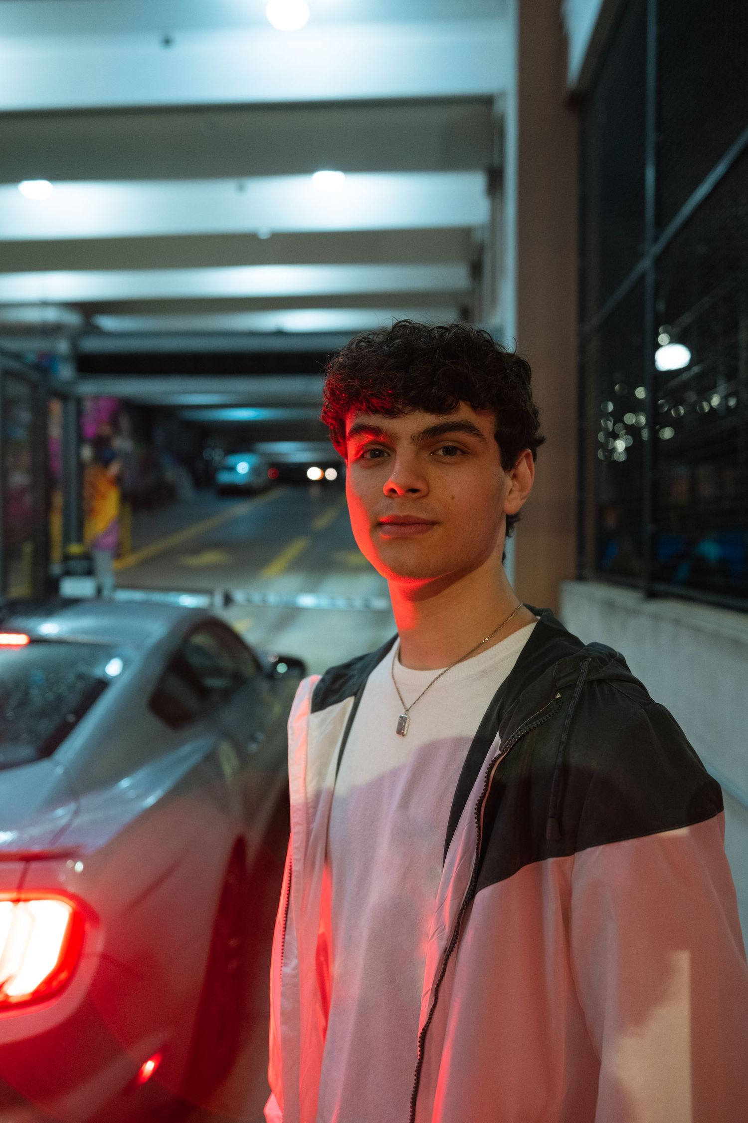 Young man with curly hair wearing a colorful jacket stands in a parking garage at night, lit by red car lights.