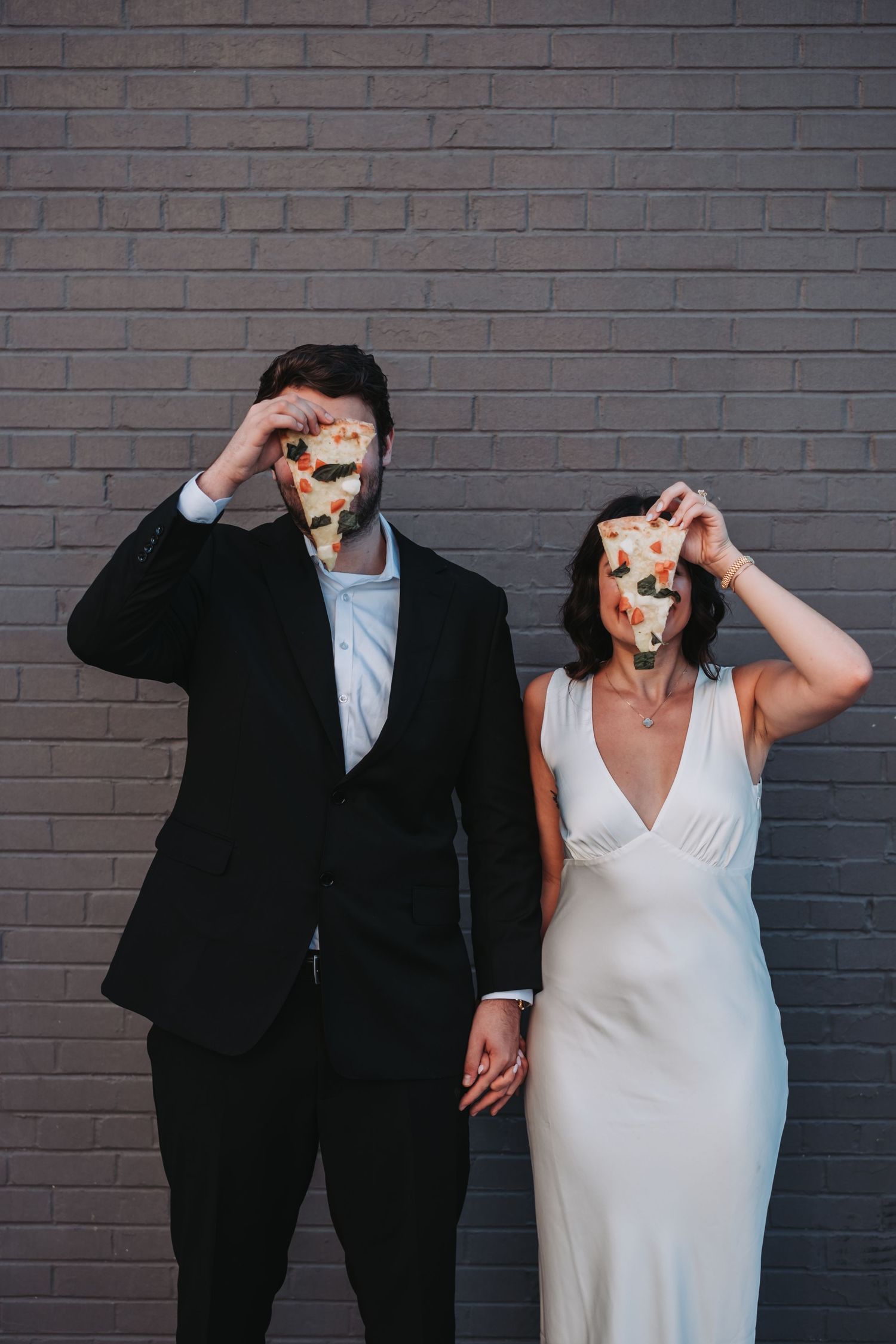 Couple in formal attire holding donuts in front of their faces against brick wall.