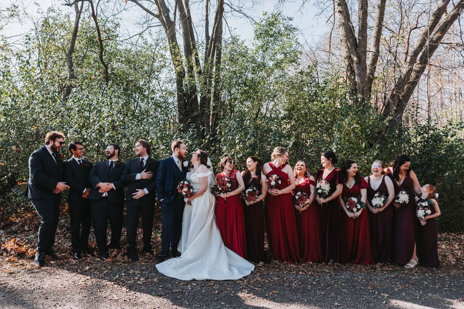 Wedding party in burgundy and black attire posing outdoors among bare trees.