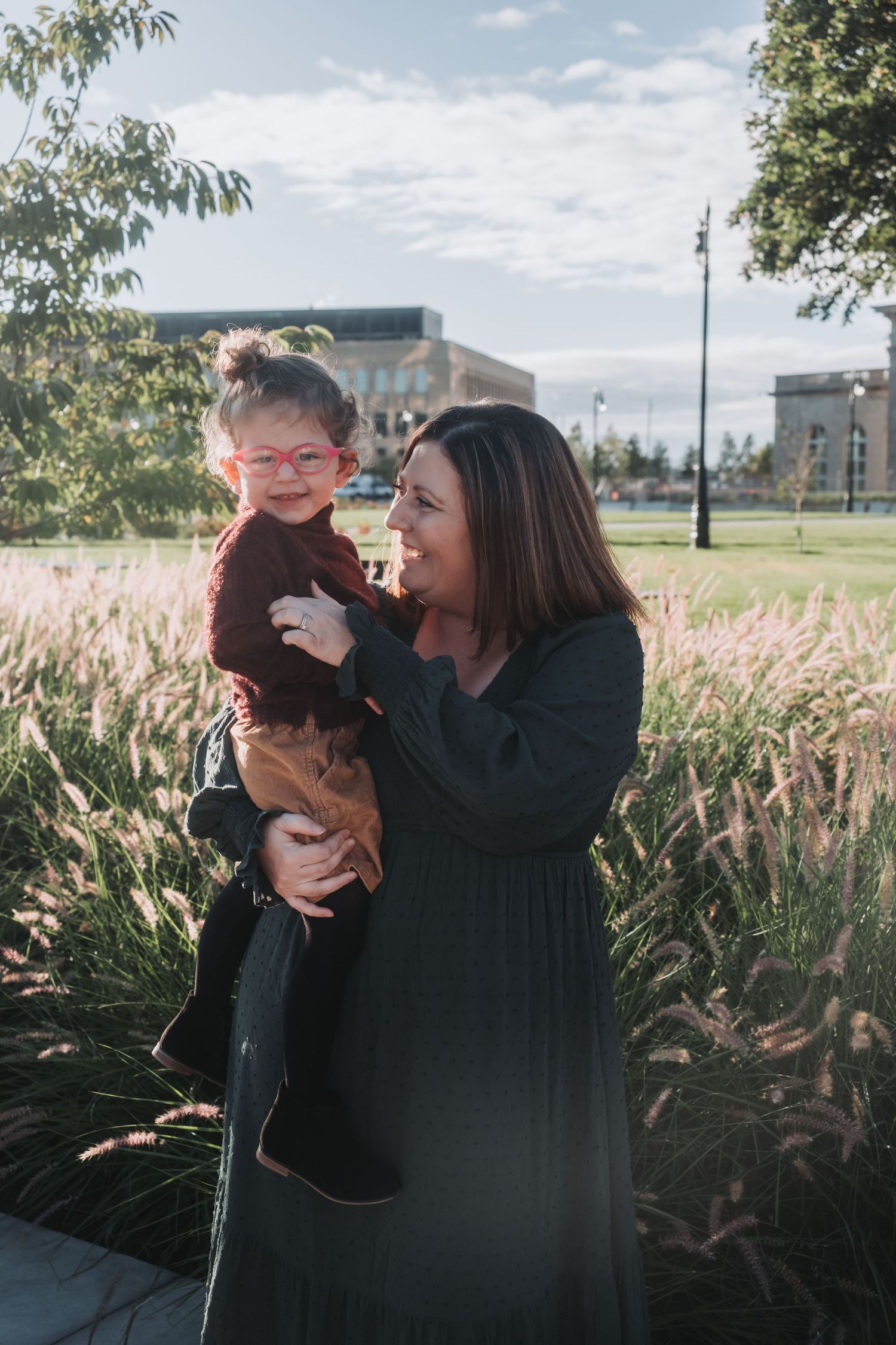 A woman in a green dress holds a smiling toddler in pink glasses outdoors near tall grasses.
