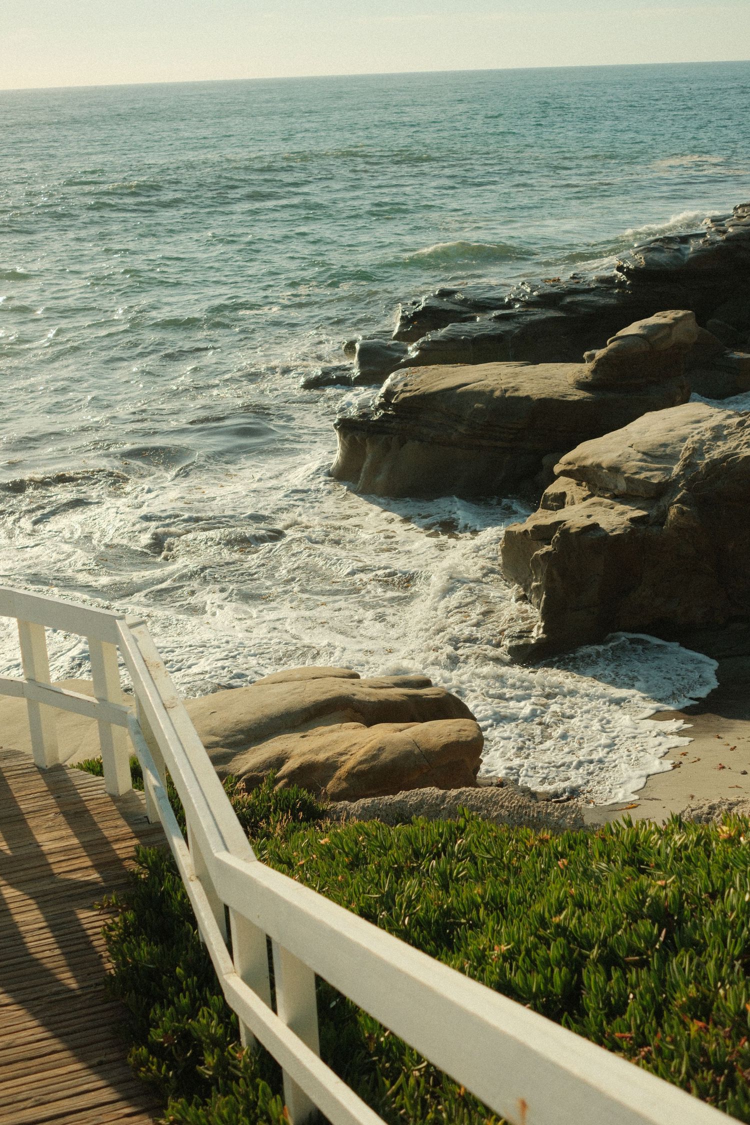 Wooden boardwalk with white railing leading to rocky ocean coastline with waves crashing against boulders.