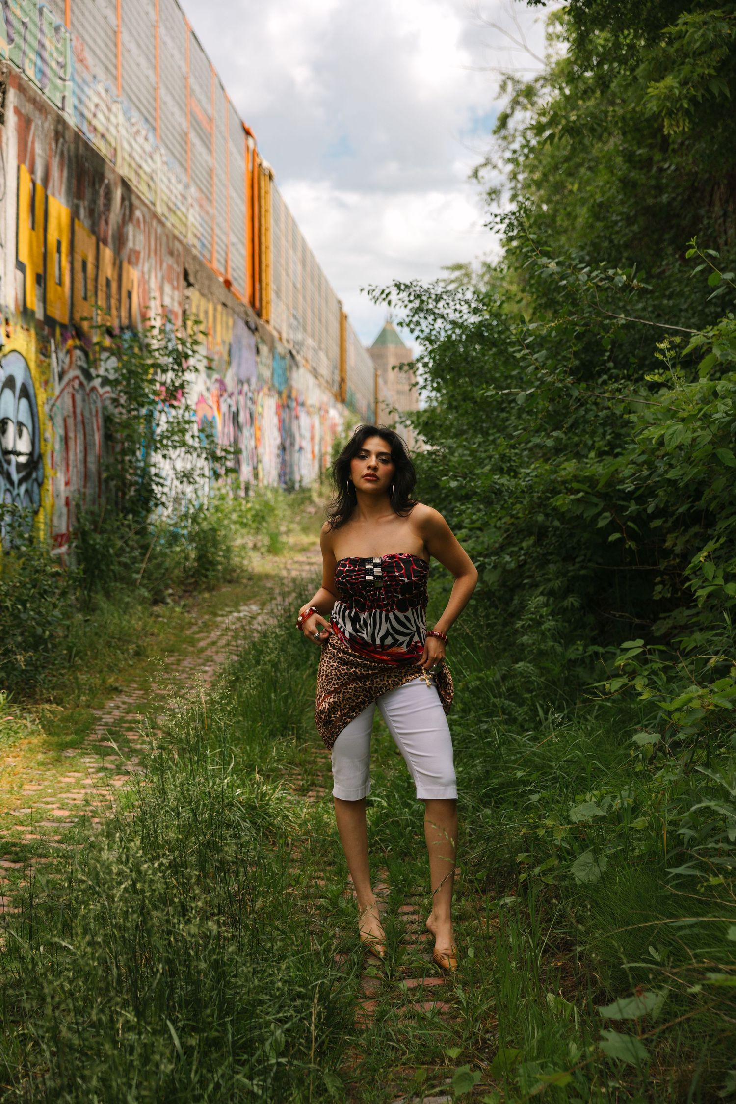 Young woman in strapless top and white capris poses barefoot in an urban alley with colorful graffiti walls.
