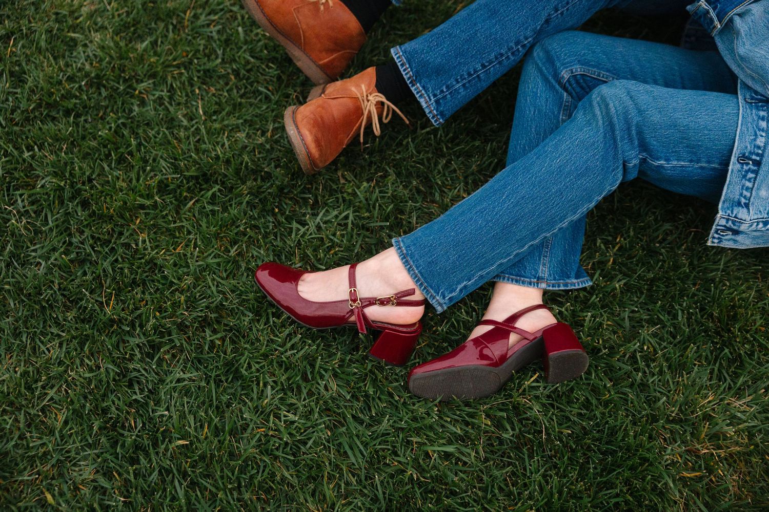 Close-up of a woman's red Mary Jane heels and man's brown boots on lush green grass.