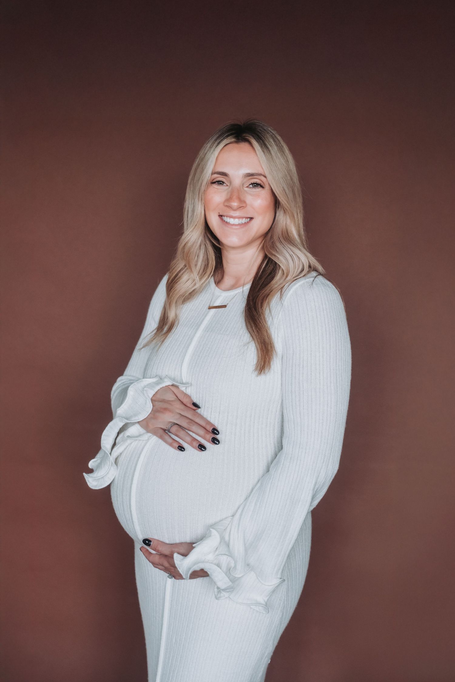 Smiling pregnant woman in white ribbed dress cradling baby bump against brown background.