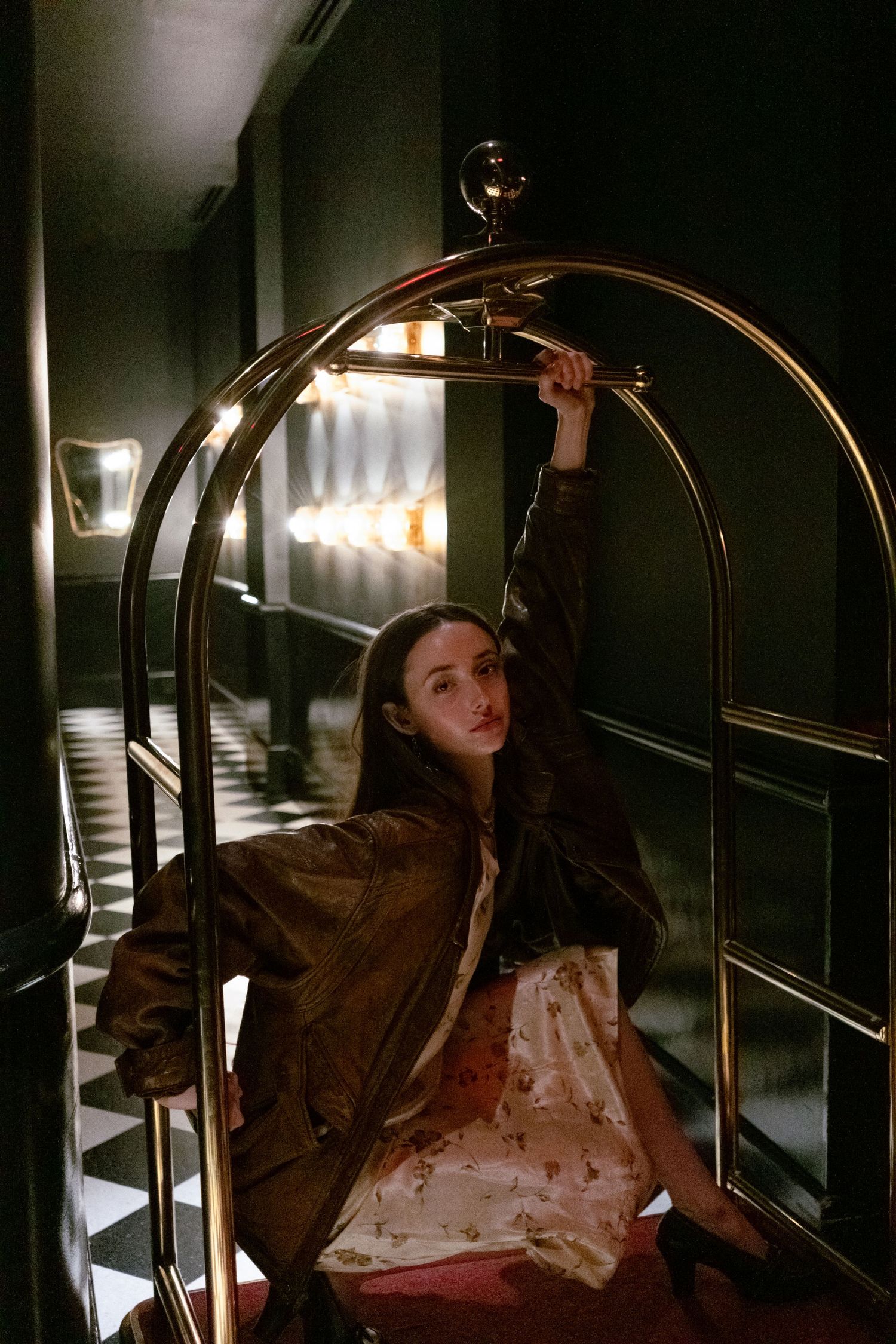 Young woman in brown jacket and floral dress posing on a gold luggage cart in a dimly lit hotel hallway.