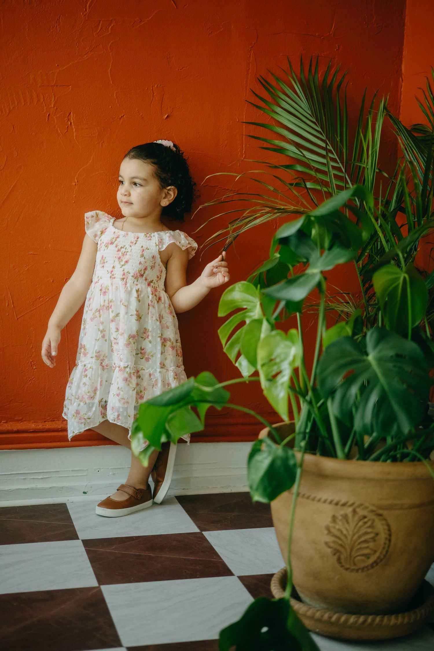 Young girl in floral dress standing by red wall with tropical potted plants on checkered floor.
