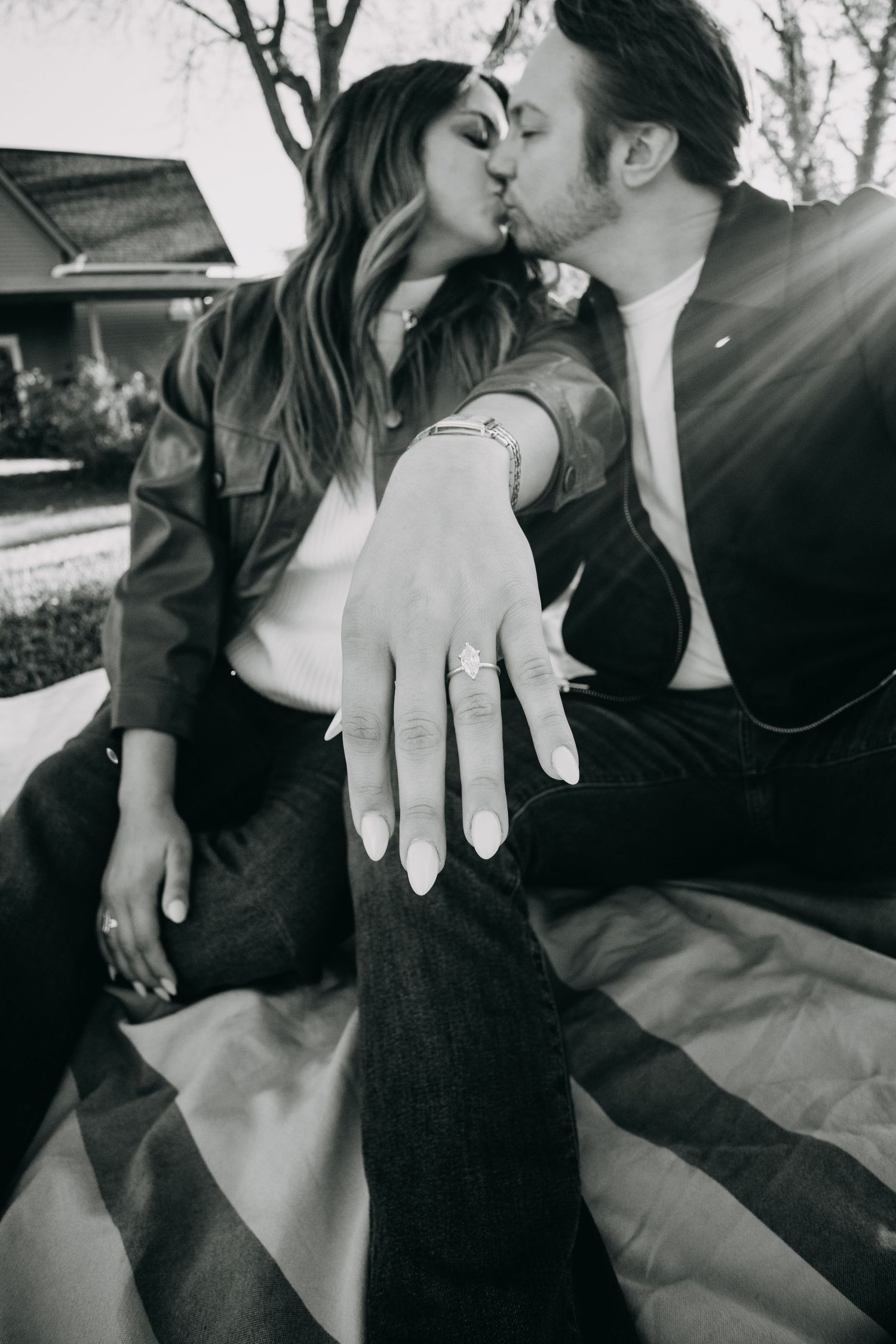 Black and white photo of an engaged couple kissing while showing off a diamond engagement ring outdoors.