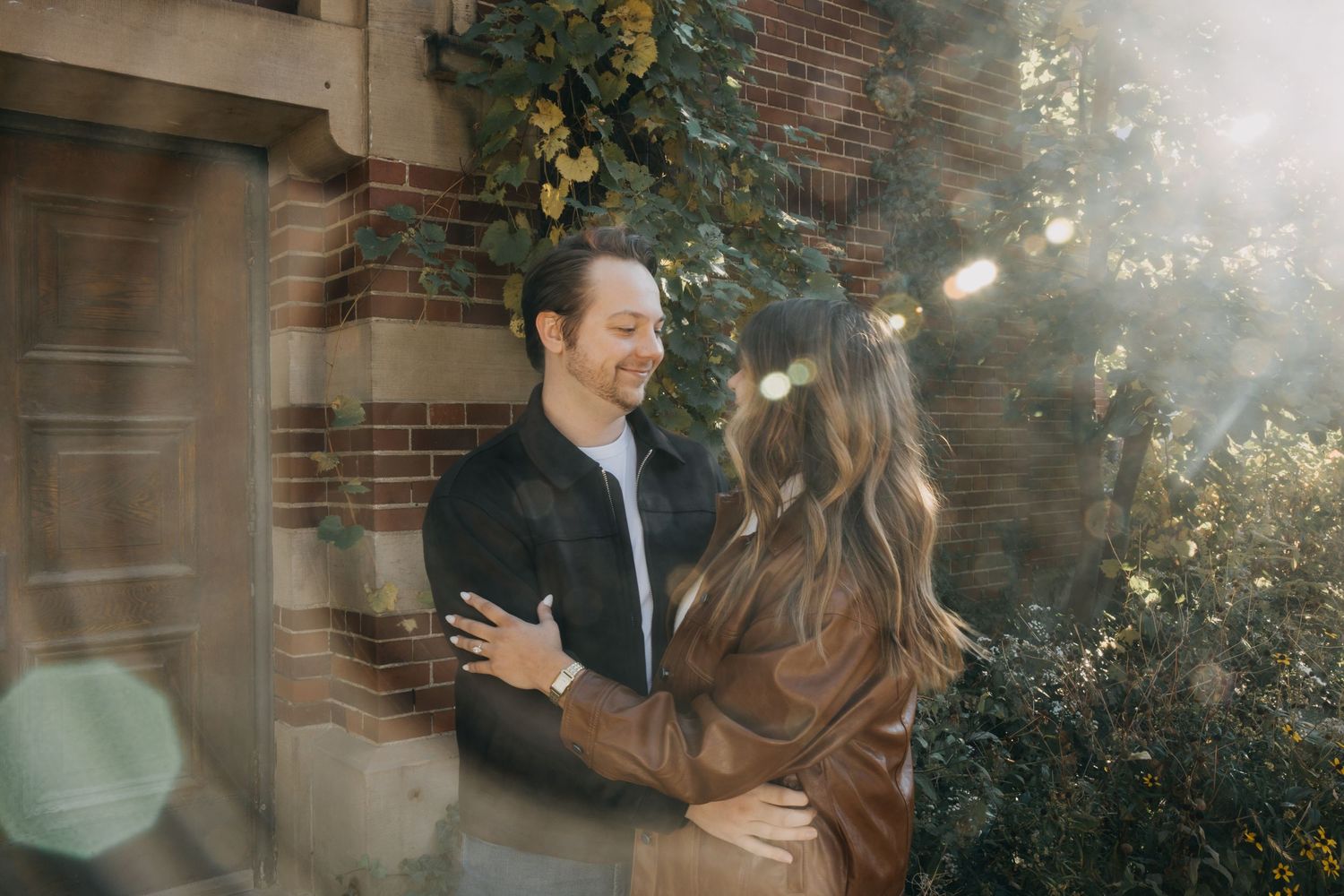 A couple shares a romantic moment outdoors, embracing near a brick wall with lush greenery in warm sunlight.