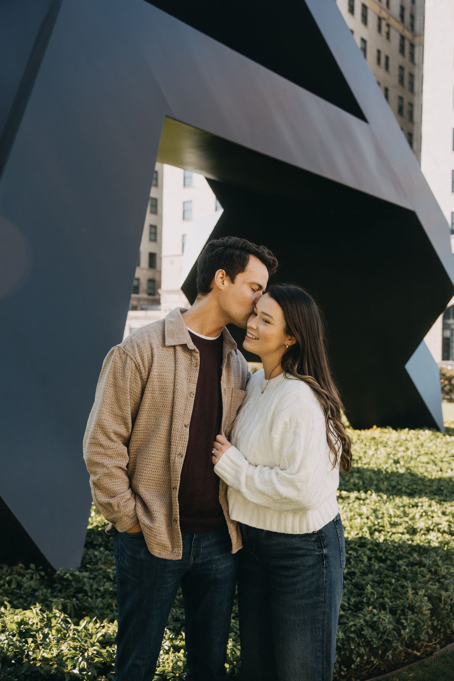 A couple shares tender moments under a black umbrella against an urban backdrop with greenery.