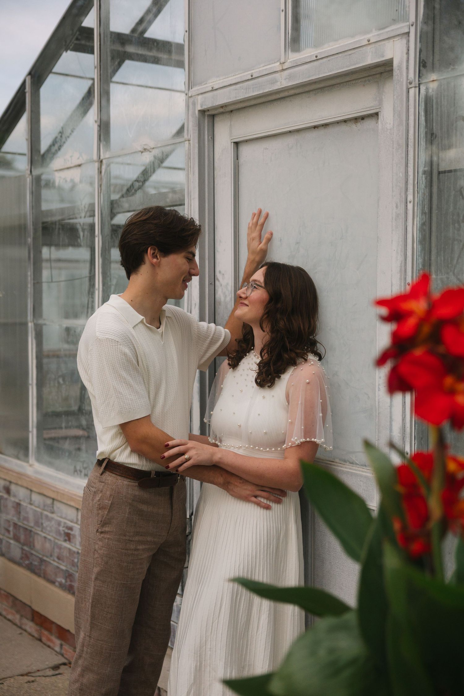 Romantic couple posing near greenhouse windows with red flowers, woman in white dress, man in casual attire.