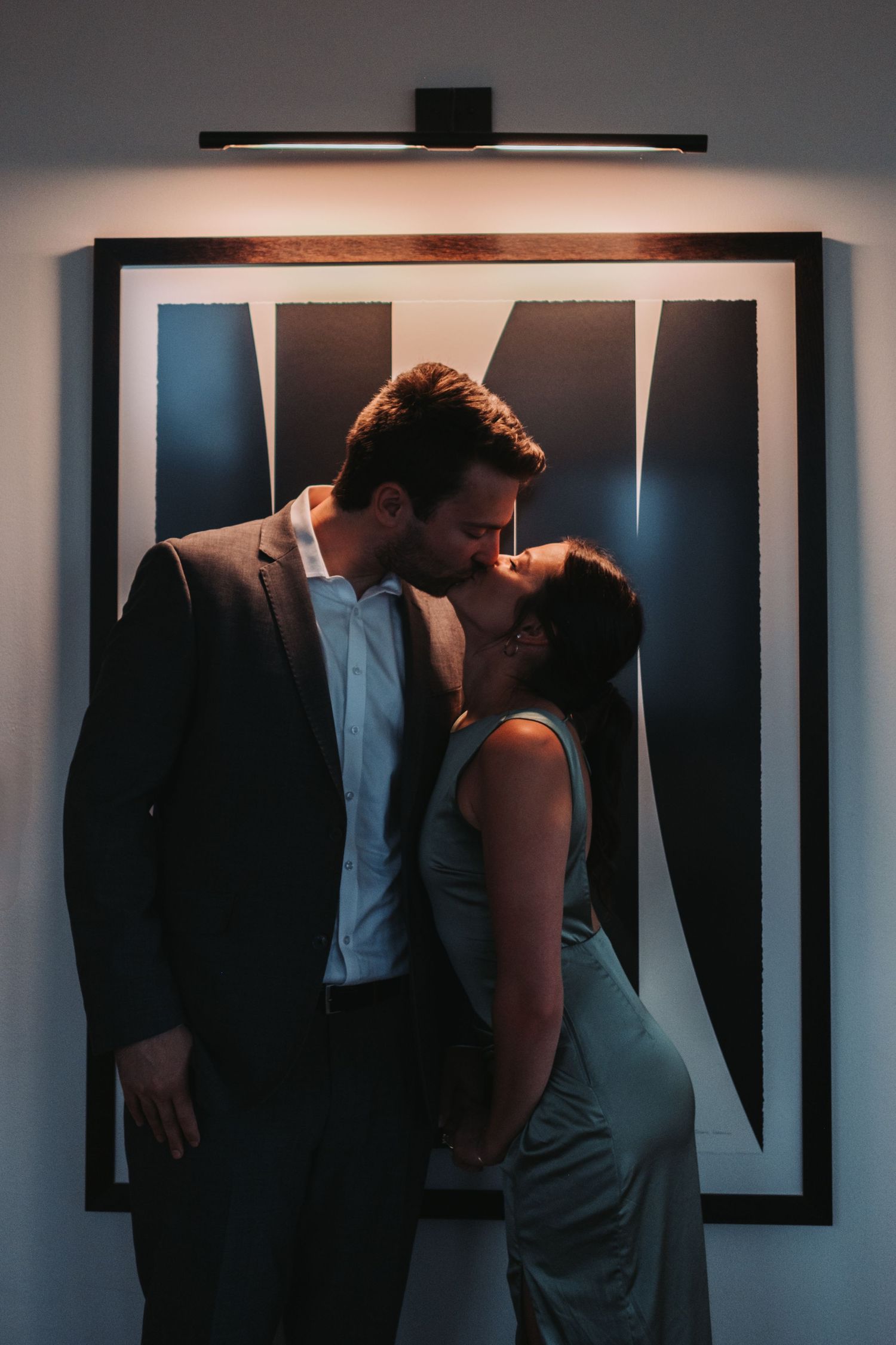 A couple shares a romantic kiss in front of a framed black and white artwork, lit by a warm overhead light.