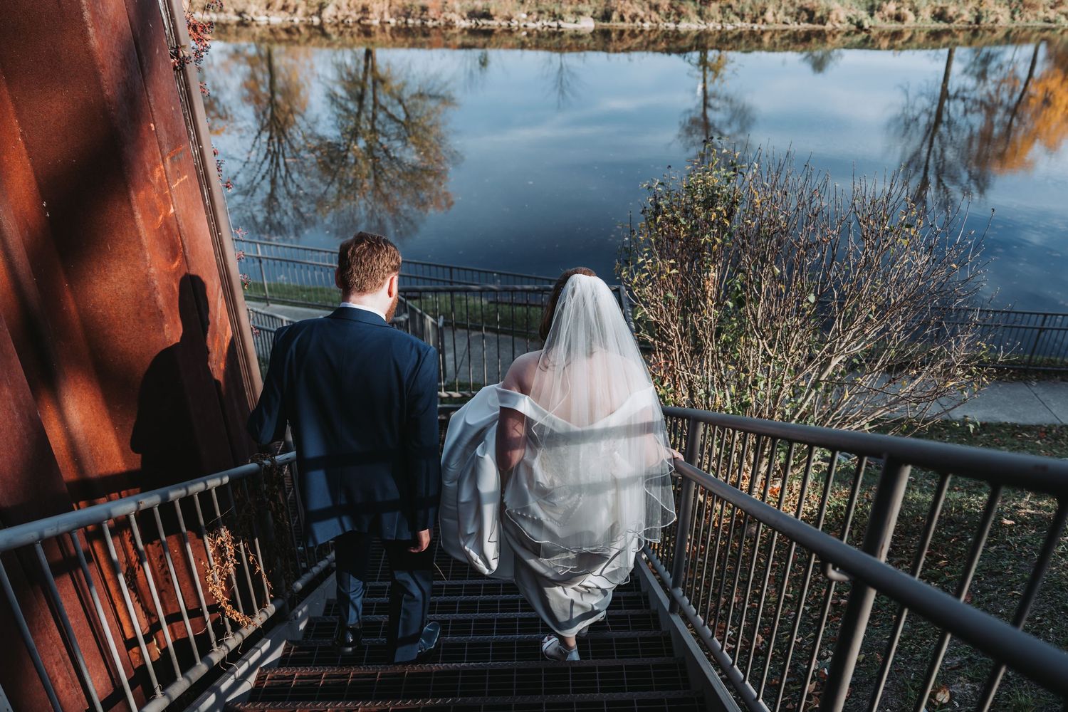 A wedding couple descends outdoor stairs overlooking a reflective lake at dusk.