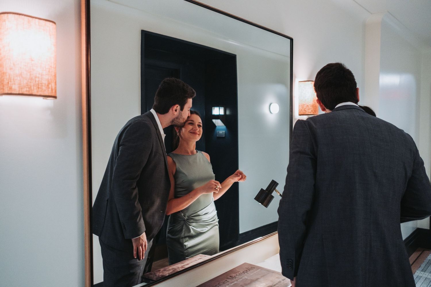 Couple sharing a romantic moment in front of a large mirror while a man in a suit stands nearby.
