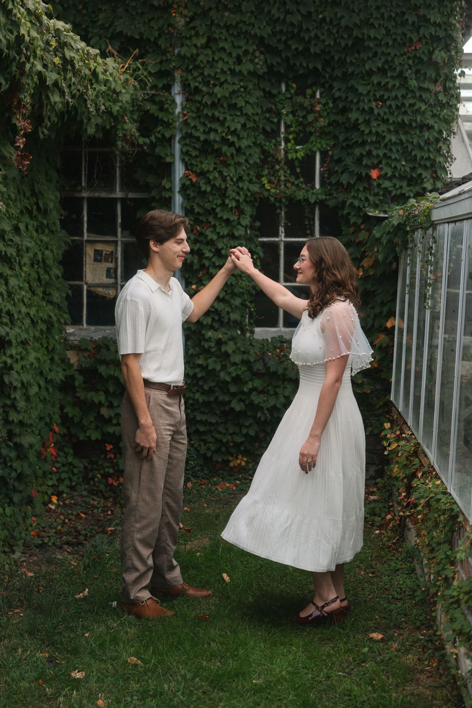 A couple dances outdoors near an ivy-covered building, woman in white dress holding hands with man in casual attire.