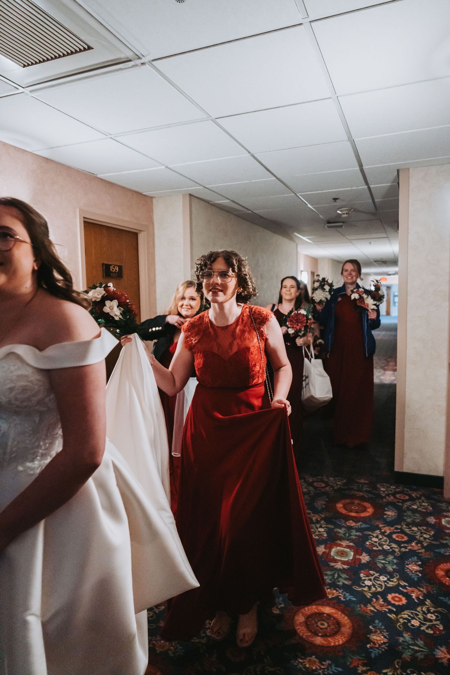Wedding party laughing while walking through hotel corridor with floral carpet.