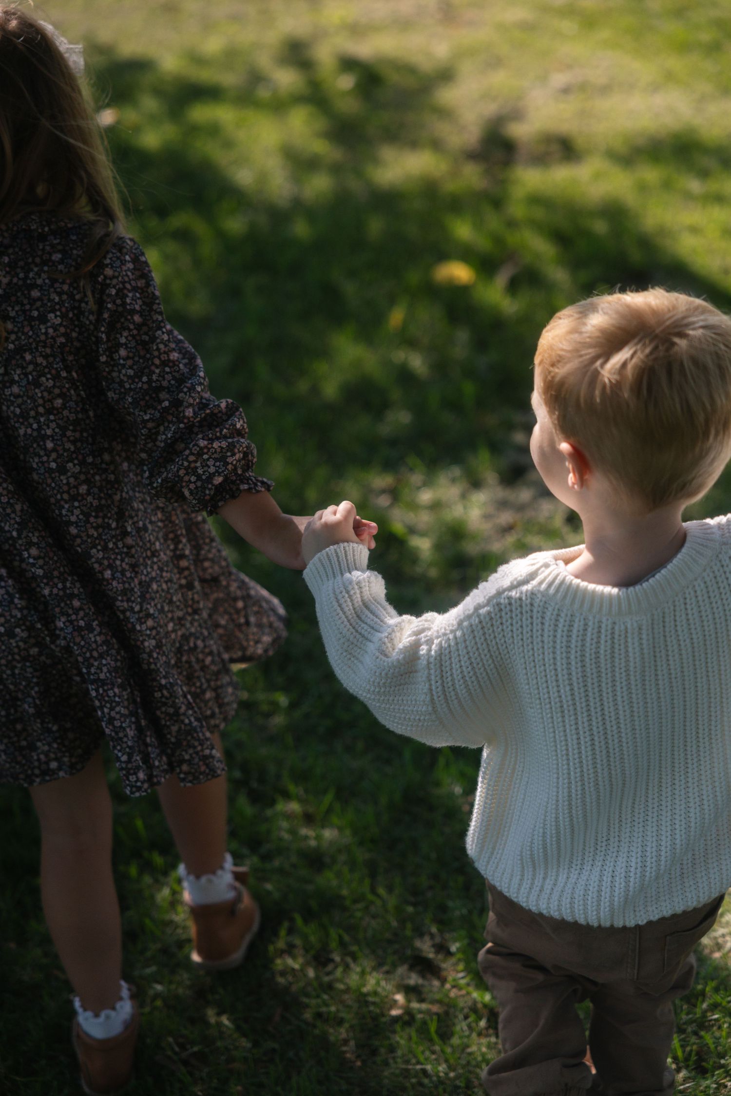 A child in a white sweater walks while holding hands with someone wearing a dark floral dress in a grassy area.