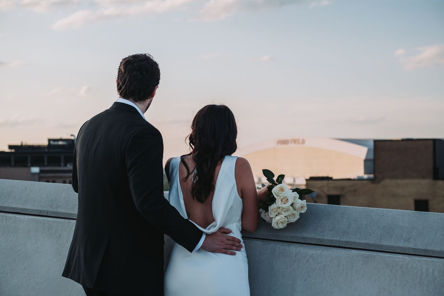 Bride and groom standing on rooftop at sunset, bride holding white roses bouquet, overlooking city skyline.
