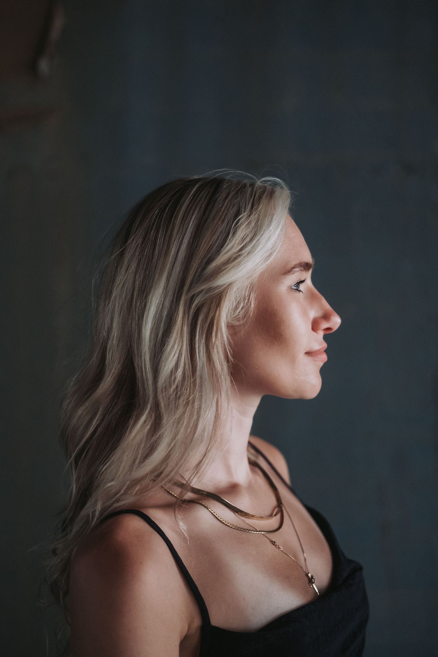 Profile portrait of a blonde woman wearing layered gold necklaces and black top against a dark gray background.