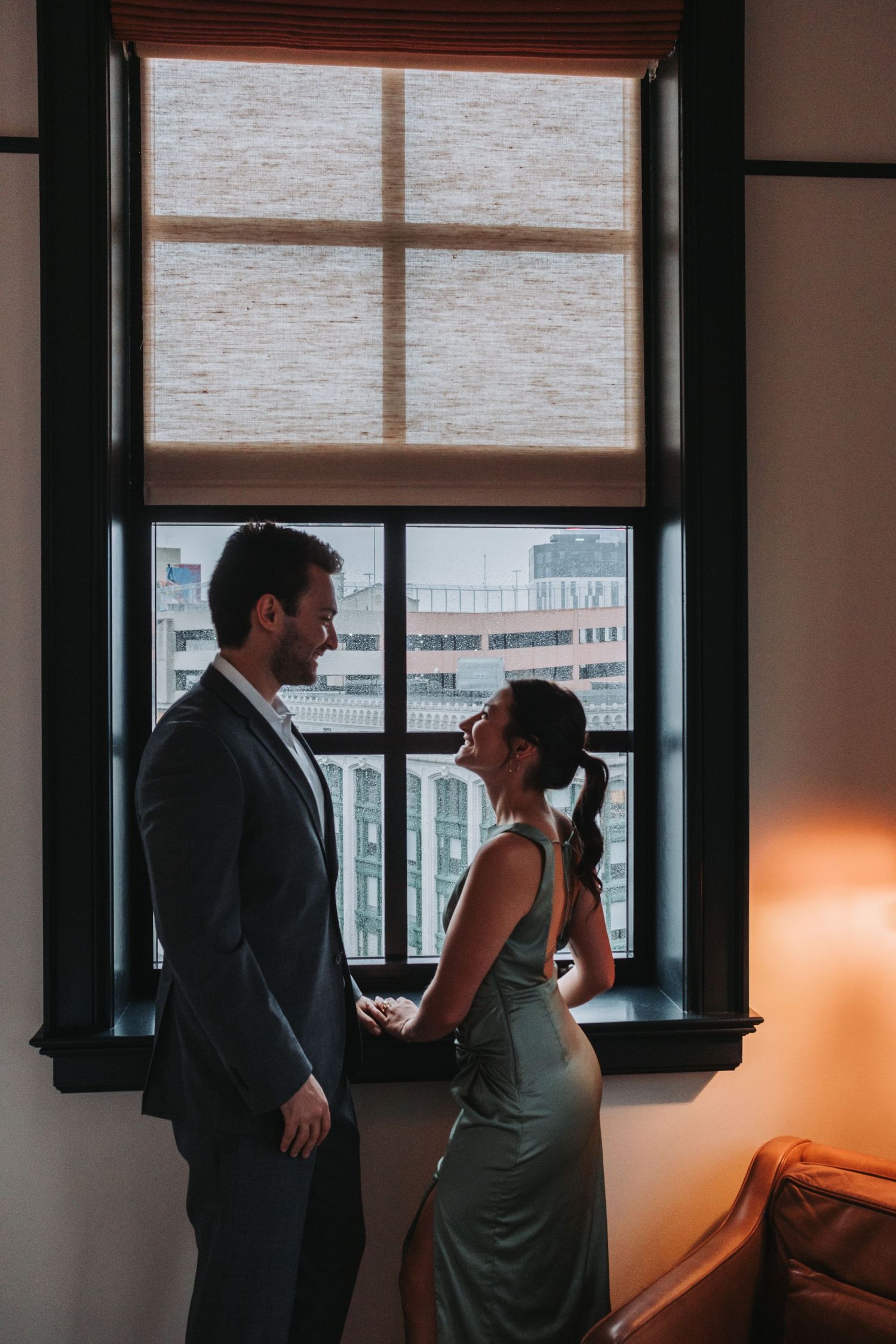 Couple holding hands and smiling by a city window, dressed in formal attire for a special occasion.