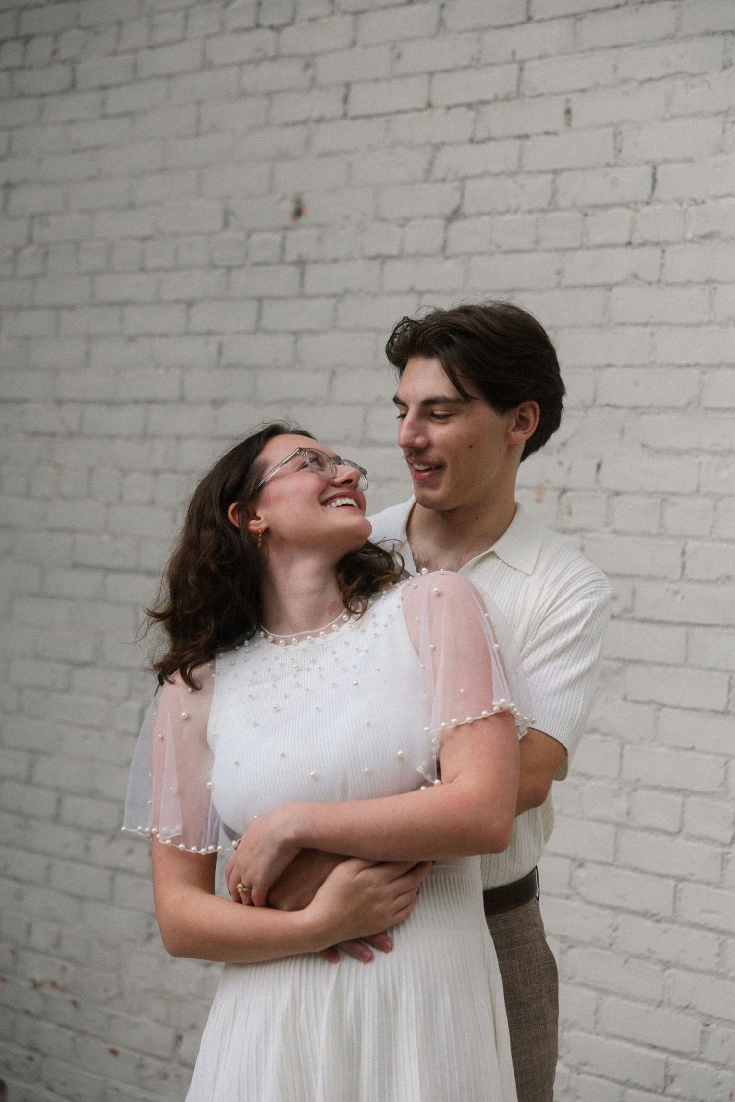 A young couple embraces and smiles at each other against a white brick wall, woman in white dress with pink sleeves.