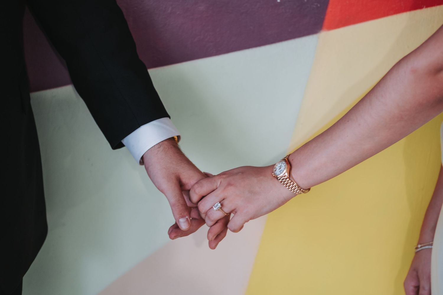 Couple holding hands with wedding ring, wearing suit and gold bracelet watch, against colorful background.