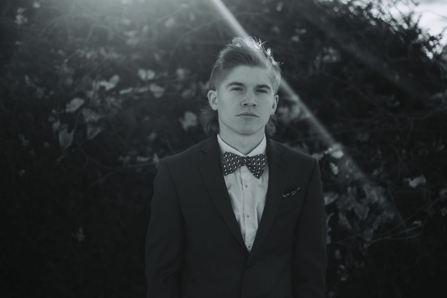 Young man in black suit and bow tie posing outdoors in black and white portrait photo.