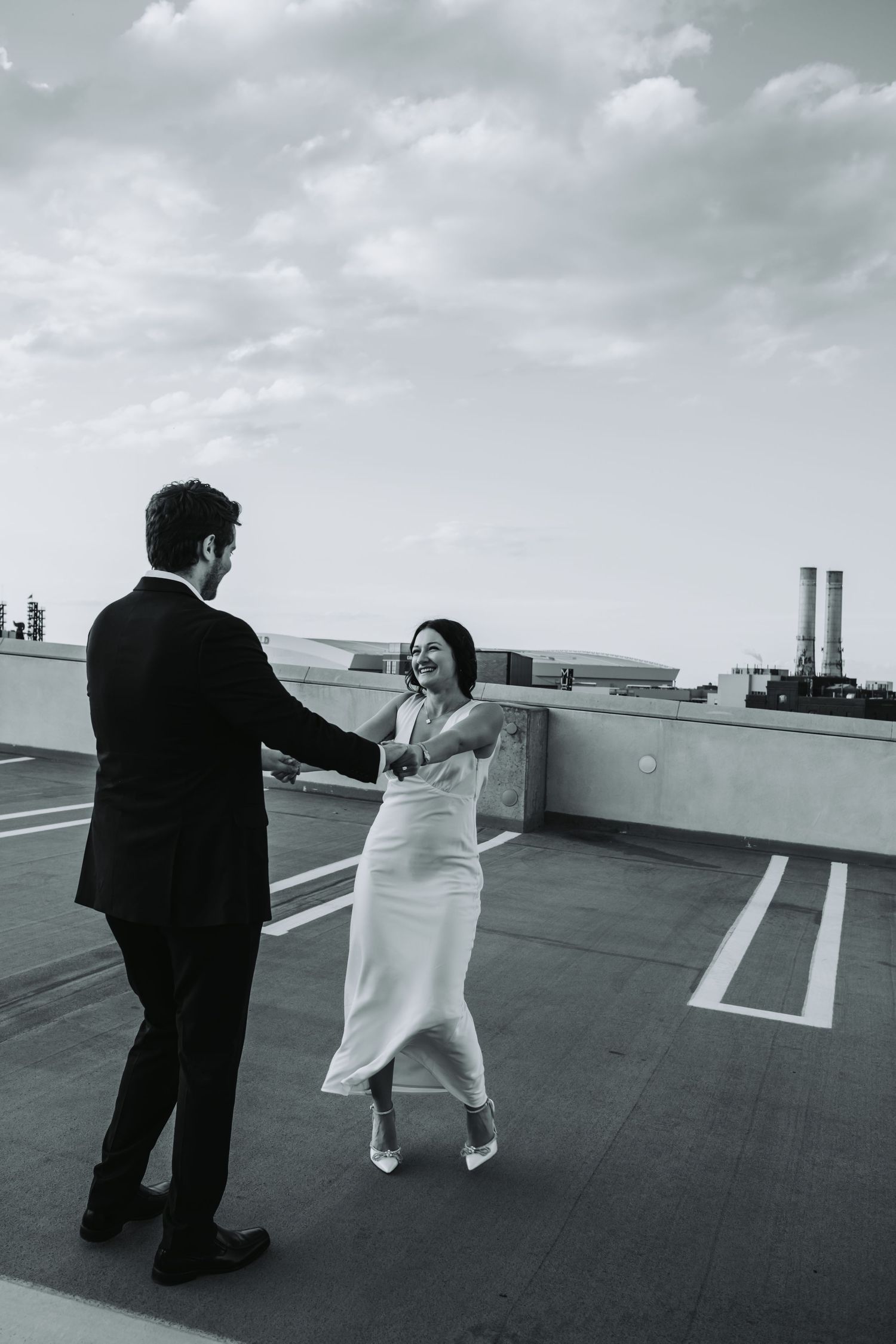 Black and white photo of a couple dancing on a rooftop, bride in white dress and groom in suit, smiling joyfully.