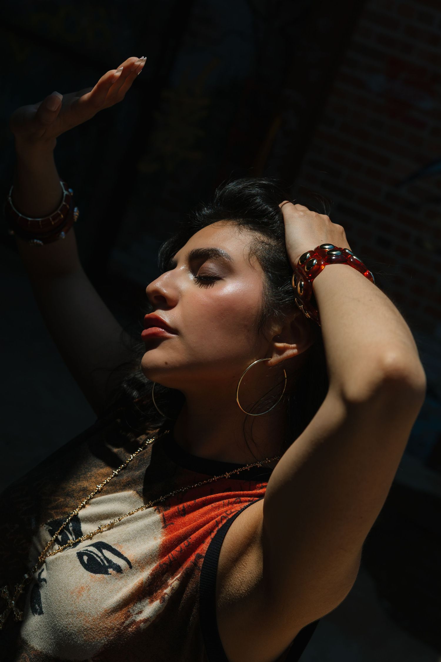 Woman with closed eyes, hoop earrings, and red bracelets posing dramatically in sunlight wearing a graphic tee.