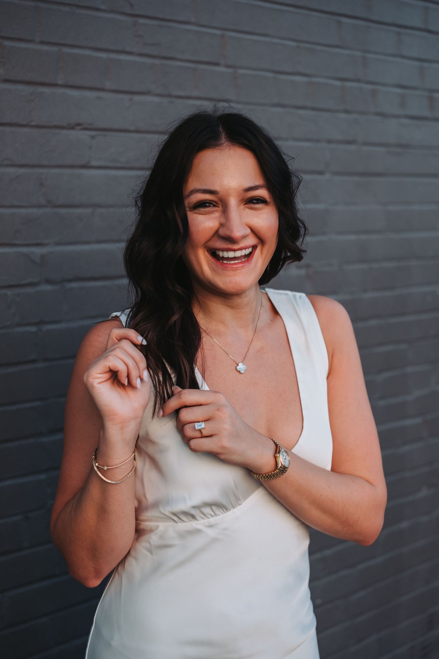 Smiling woman in white dress with necklace and watch, posing against gray brick wall.