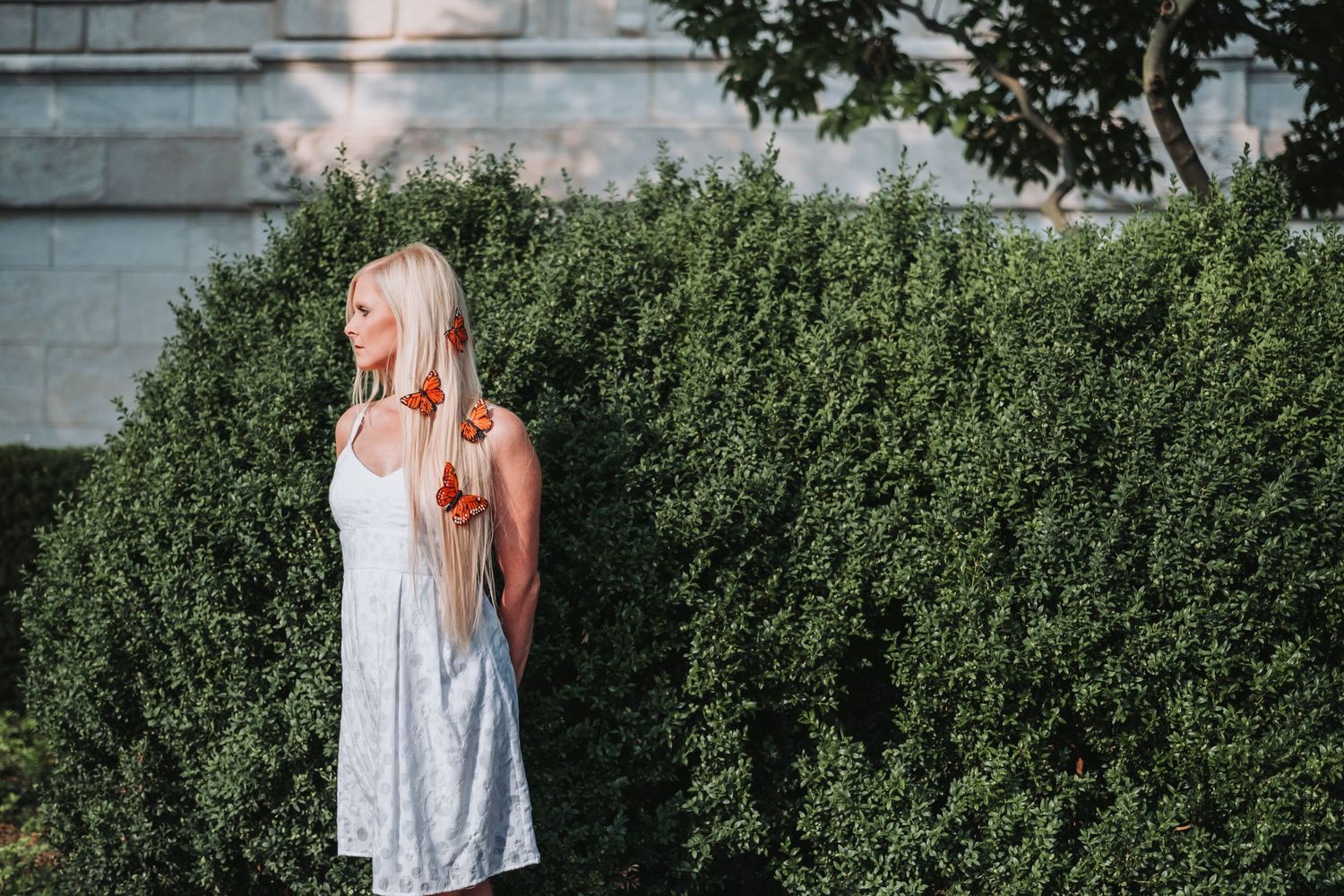 Blonde woman in white sundress standing by lush green hedges outdoors in soft evening light.