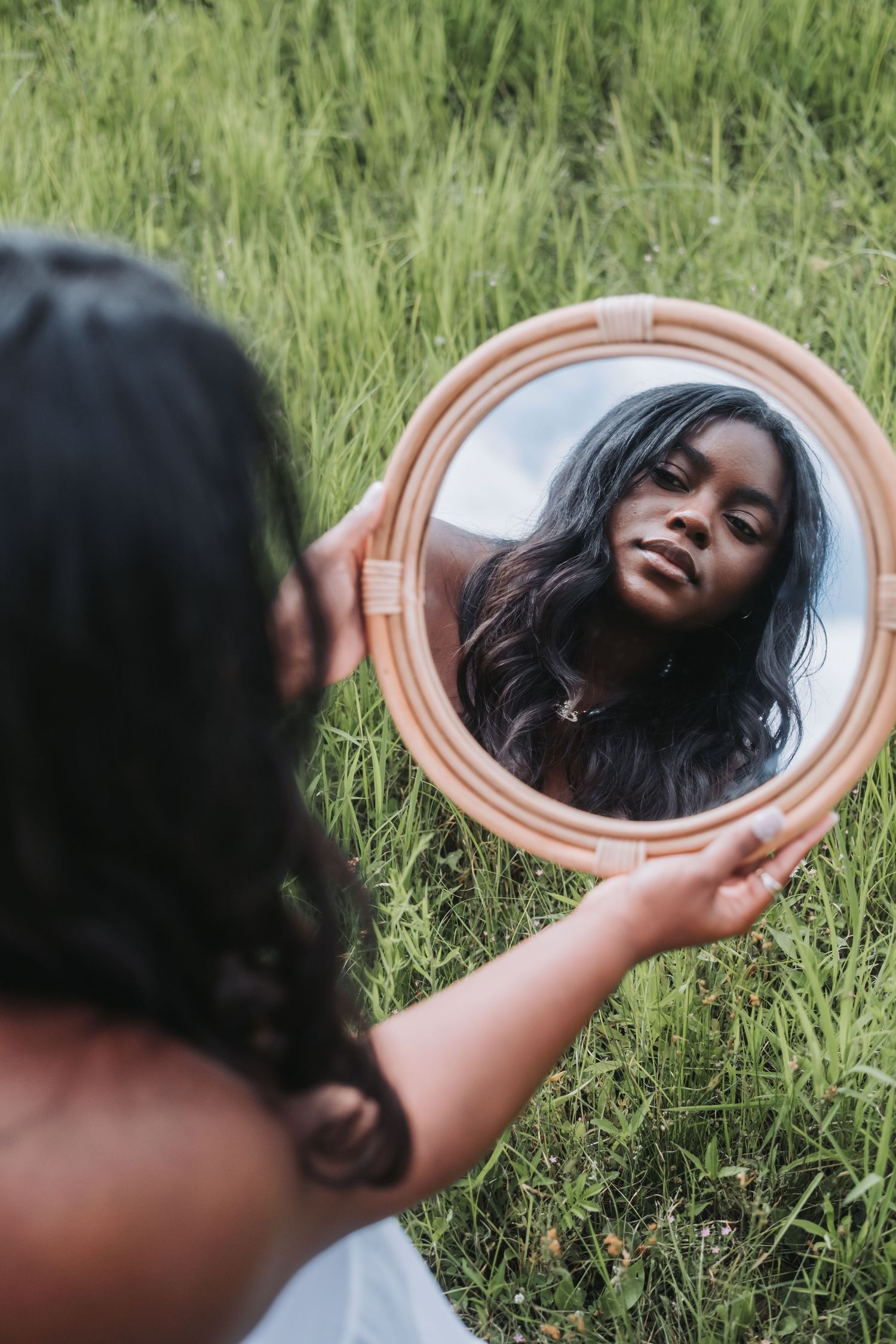 A young Black woman holds a round mirror reflecting her face while sitting in a lush green grass field outdoors.
