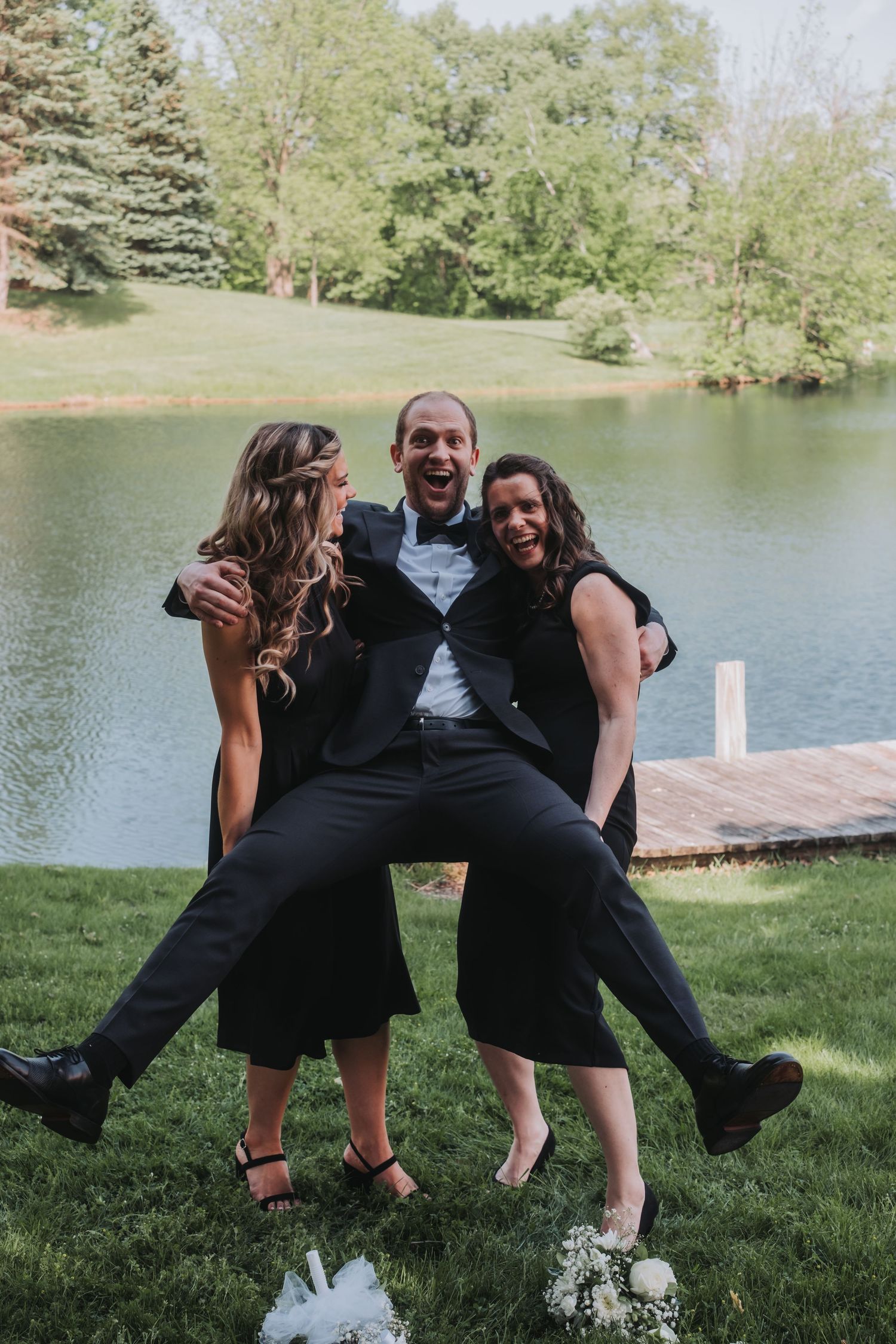 Wedding party poses playfully on grass near a lake for a fun group photo.