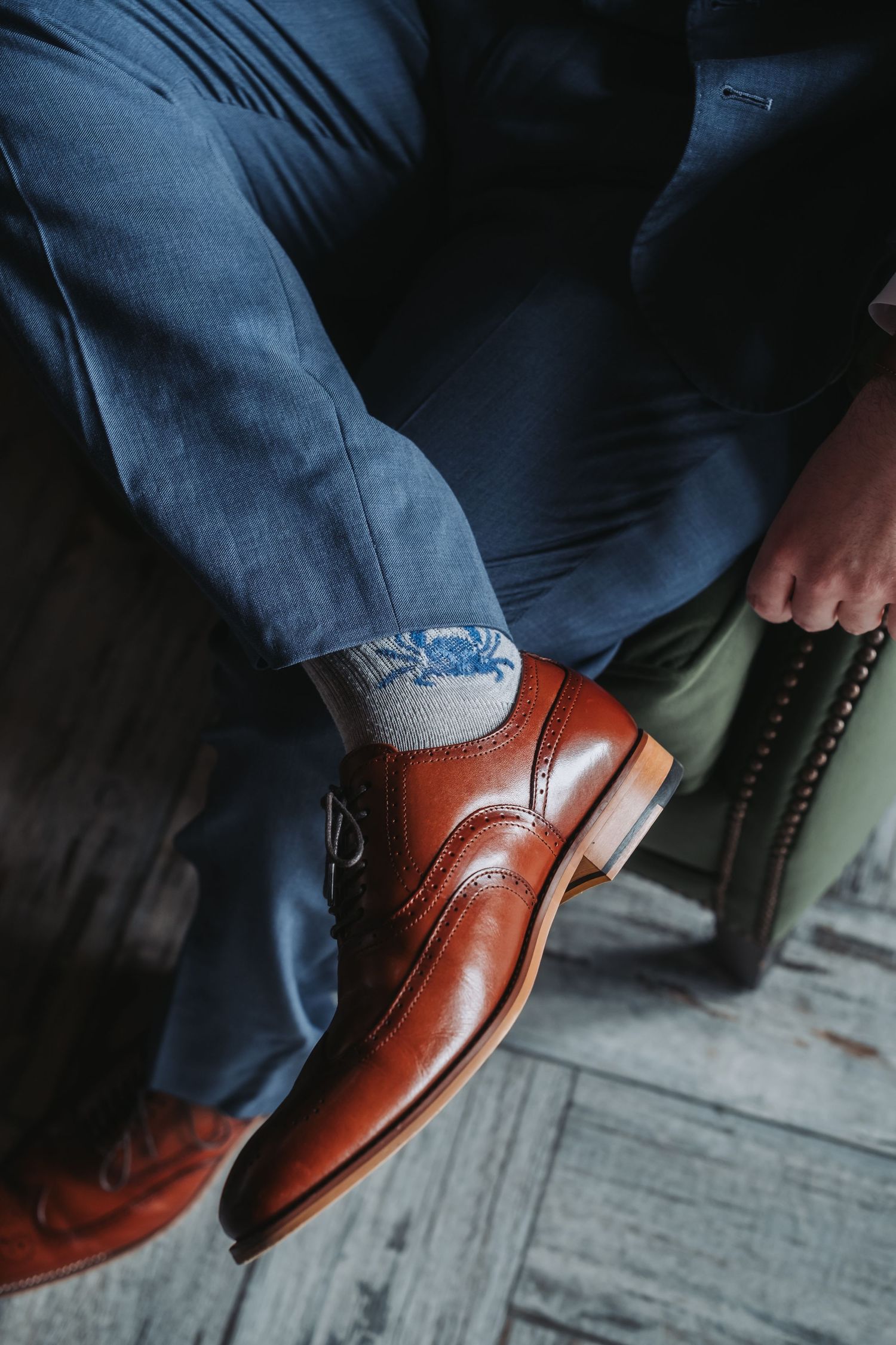 Detail shot of polished brown leather dress shoes against grey pants on wooden flooring.