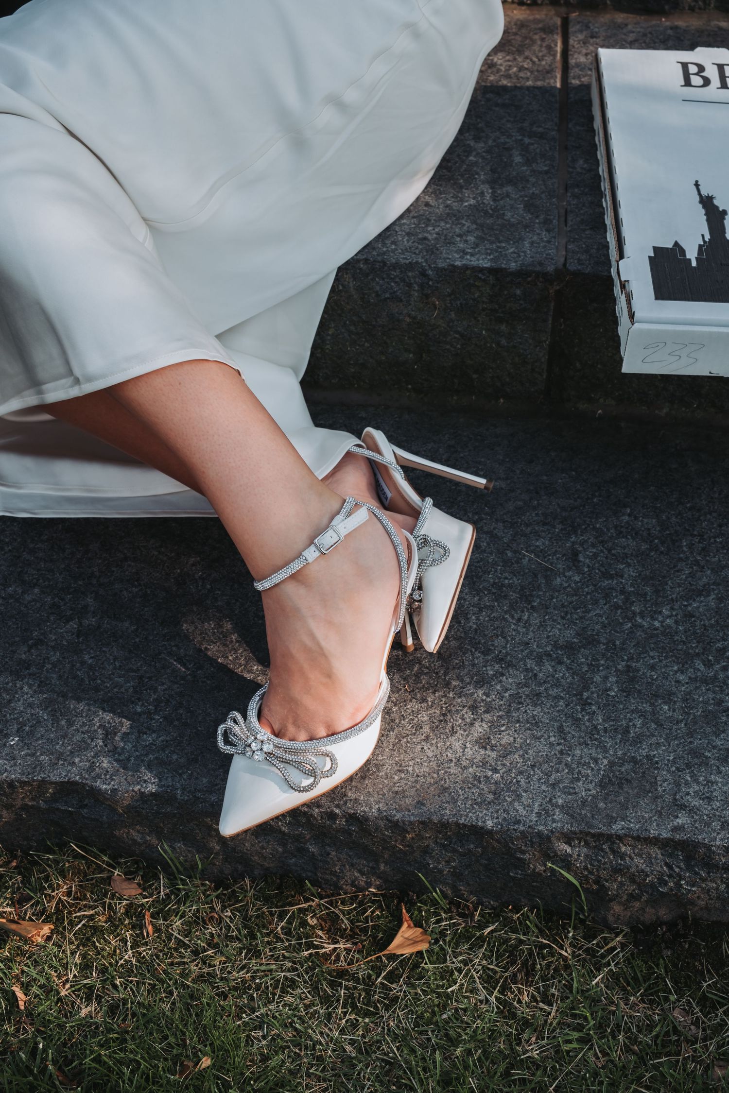 Elegant white bridal heels with crystal bow detail and ankle strap on stone steps.