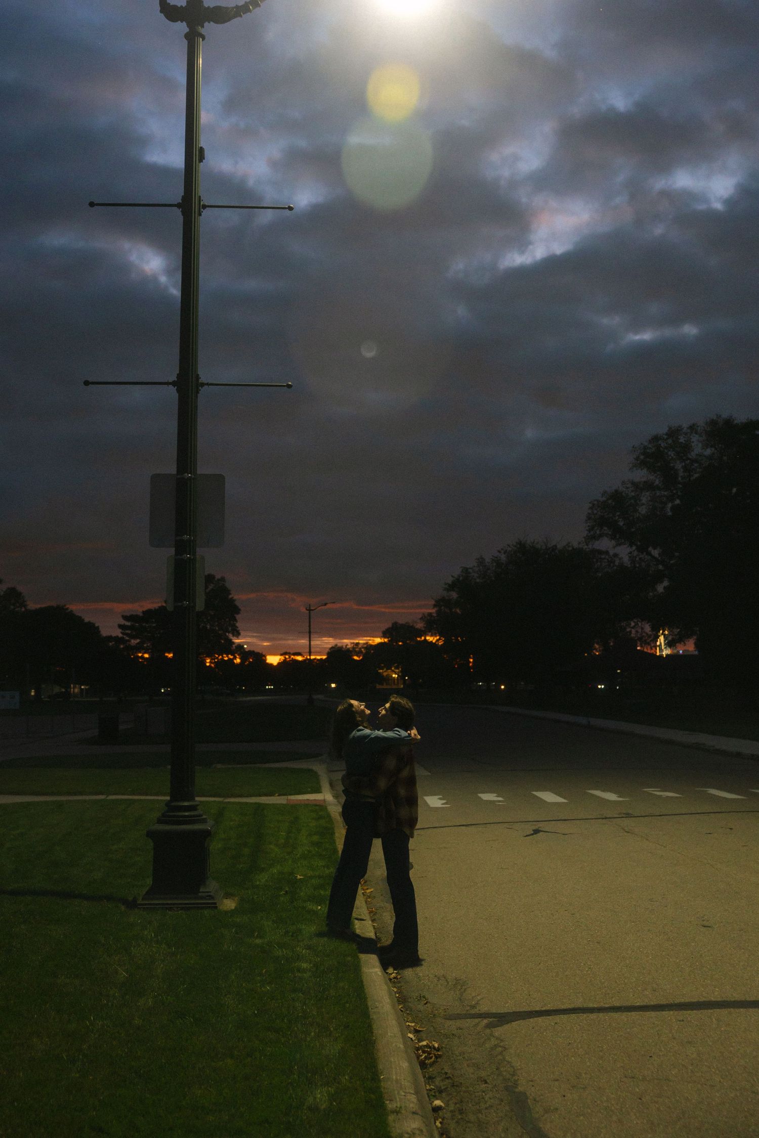 Couple embracing near a street lamp at dusk with dramatic storm clouds and colorful sunset sky in background.