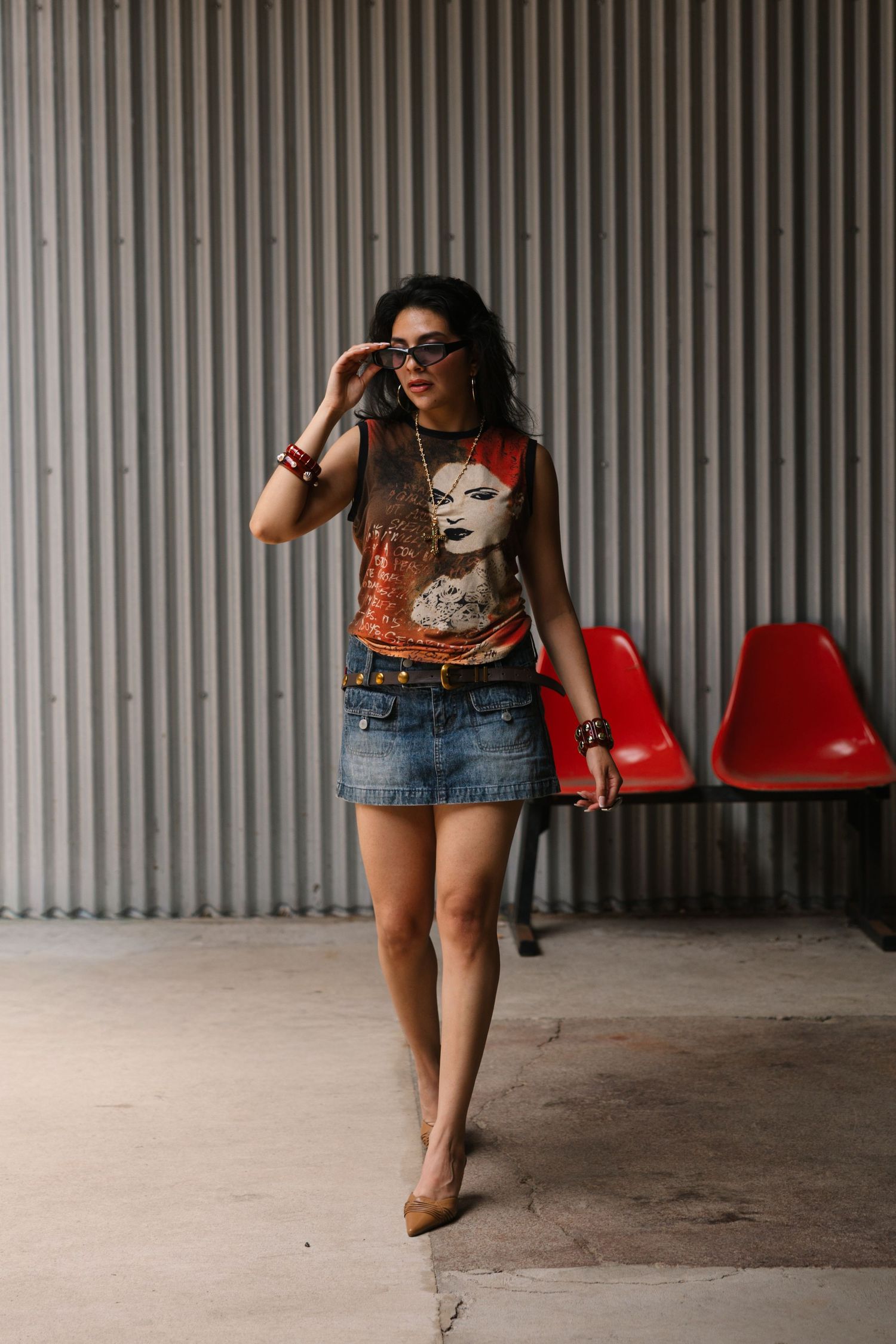 Woman in graphic tee, denim mini skirt, and sunglasses posing near red chairs against corrugated metal wall.