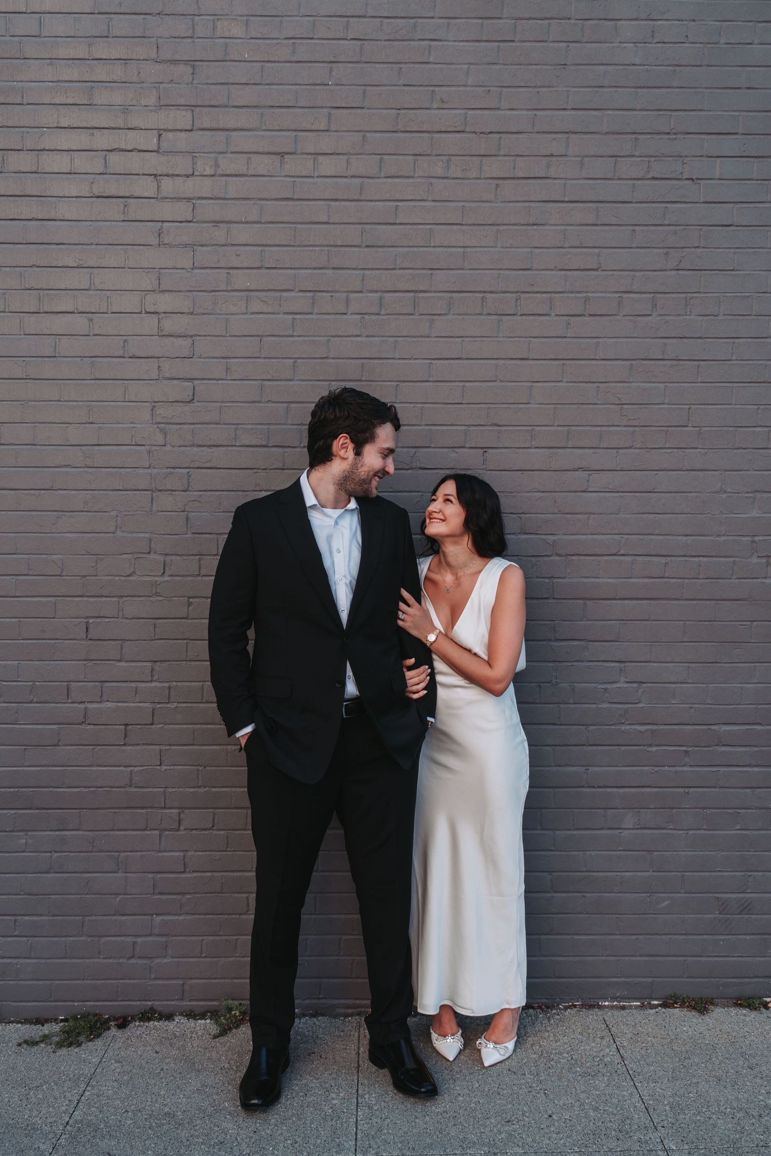 A couple in wedding attire smiling at each other against a gray brick wall, bride in white dress, groom in black suit.