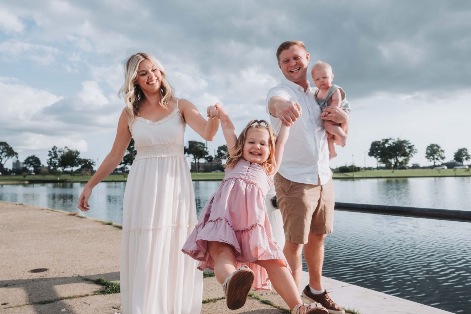 Happy family of four posing by a lake, mom in white dress, dad holding baby, daughter in pink dress.