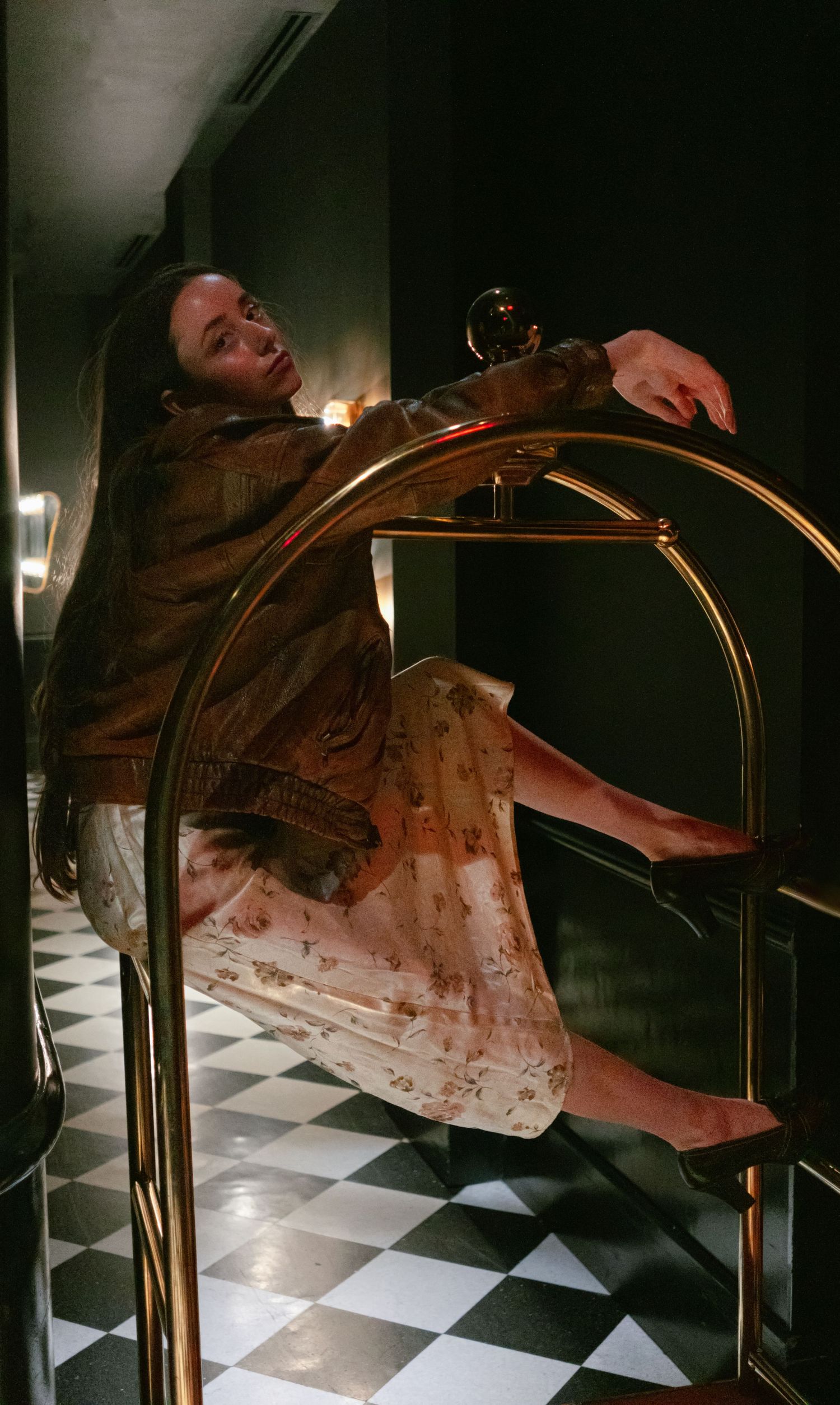 Woman in floral skirt and brown jacket draped over a gold luggage rack on a checkered floor.